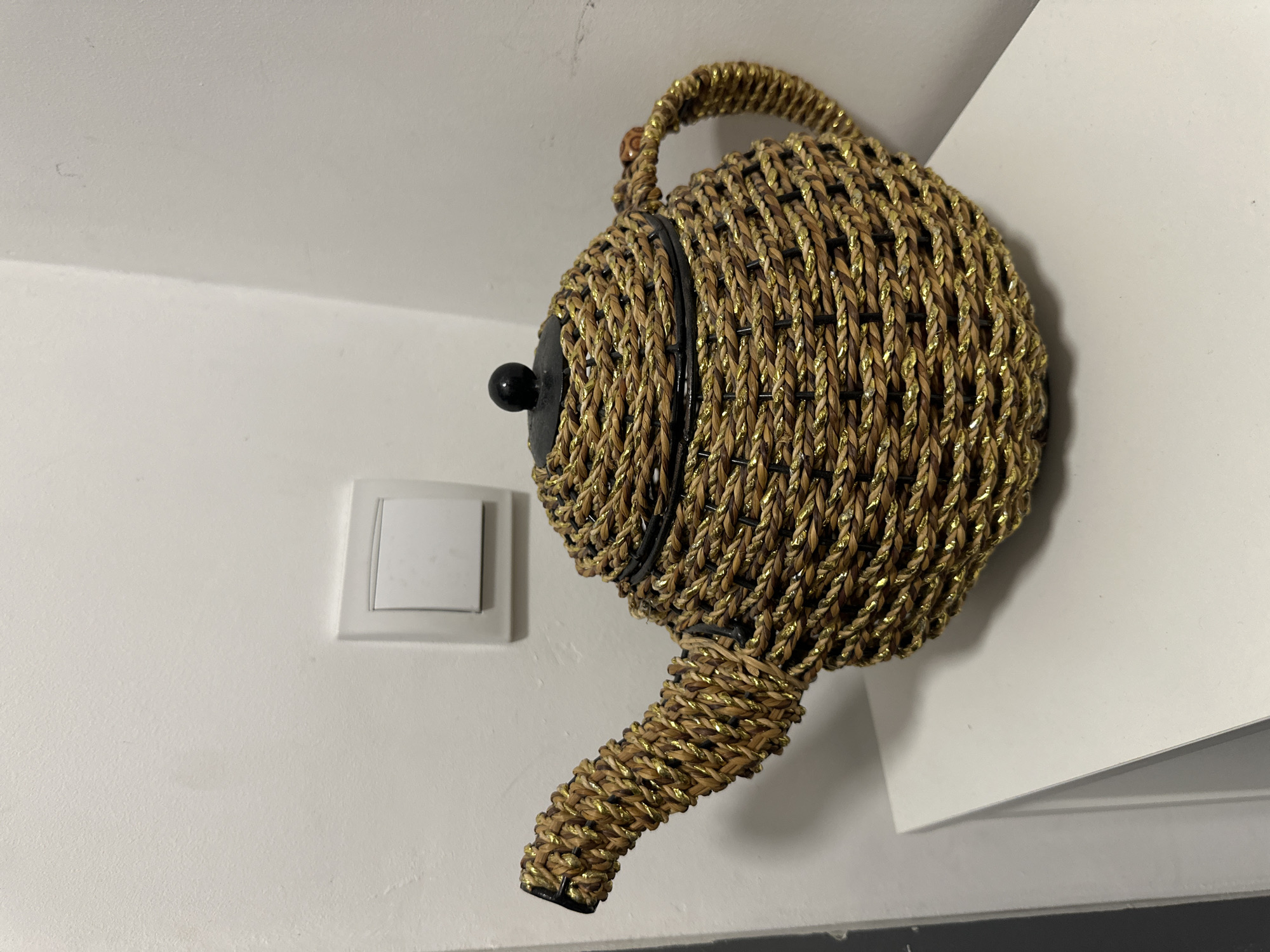 A woven teapot, made of natural fibers with metallic gold threads, rests on a white surface, likely a shelf or table. The teapot has a spout and a handle, and a black lid with a small black knob. It is positioned in a corner where two white walls meet. A white electrical outlet with a switch is visible on the wall behind the teapot. The lighting in the scene is soft, suggesting indoor lighting. The scene appears to be an interior domestic setting. This image is not tagged with a specific location in Sofia, Bulgaria.