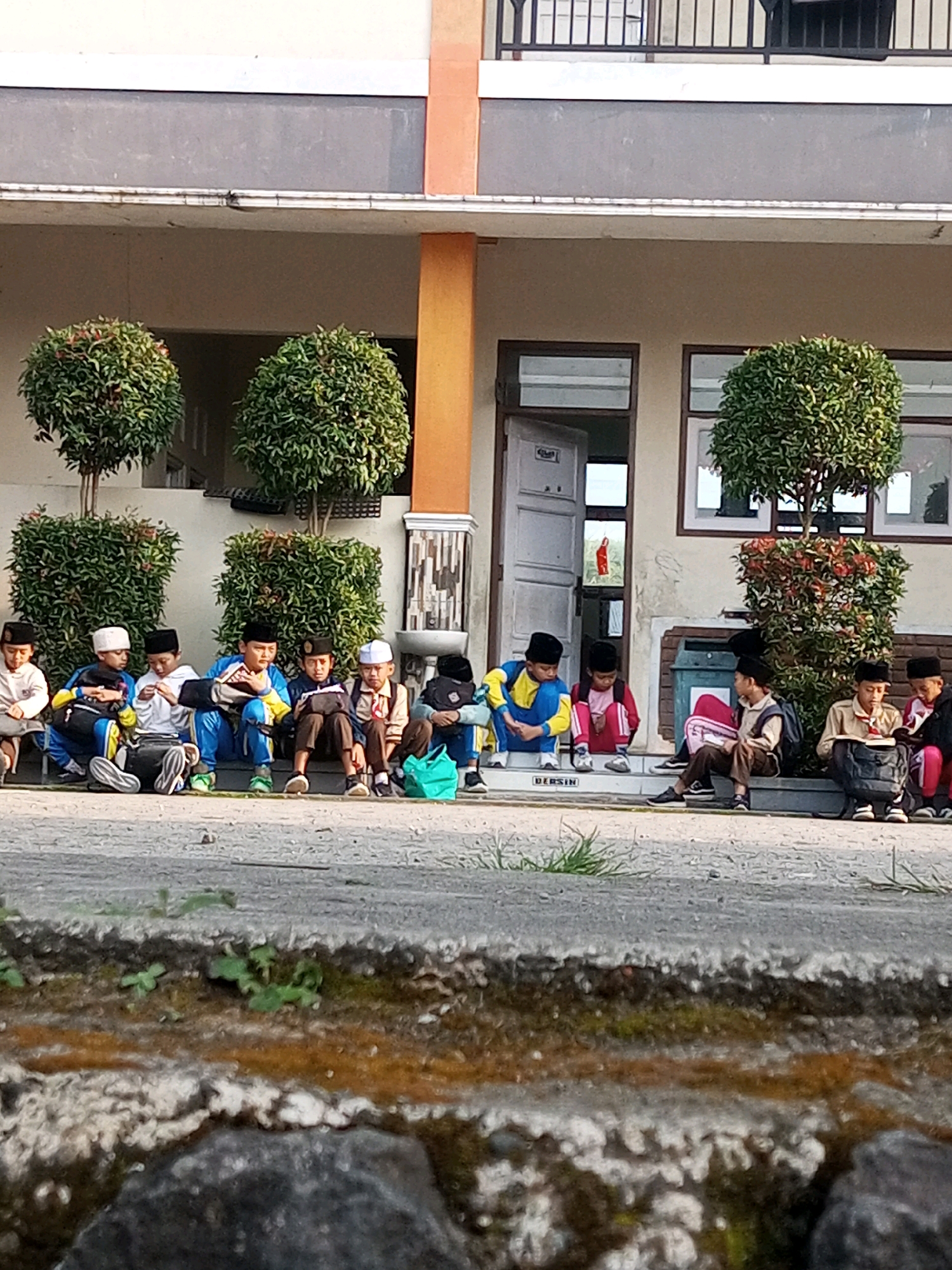 The image shows a group of young students, likely boys, sitting on a ledge in front of a building in Kalibeber, Indonesia. They are all wearing traditional Islamic caps, known as peci, and appear to be dressed in school uniforms. Some are wearing blue and yellow outfits, while others are in white or red. Many have backpacks with them. The students are seated in a line, facing forward, with some interacting with each other and others looking down.
The building behind them has a light-colored facade with dark gray trim above it. There are several windows visible, some with shutters. The entrance to the building is open, revealing a red figure inside, possibly a decoration or a person. Several potted plants with green foliage are strategically placed in front of the building, adding a touch of nature to the scene. The ground in front of the students is paved with a grayish material, and some grass is growing along the edges. In the foreground, there is a textured concrete barrier with some rocks and vegetation, suggesting an outdoor seating area or pathway. The lighting suggests it is daytime, likely morning or afternoon, with sunlight casting shadows.