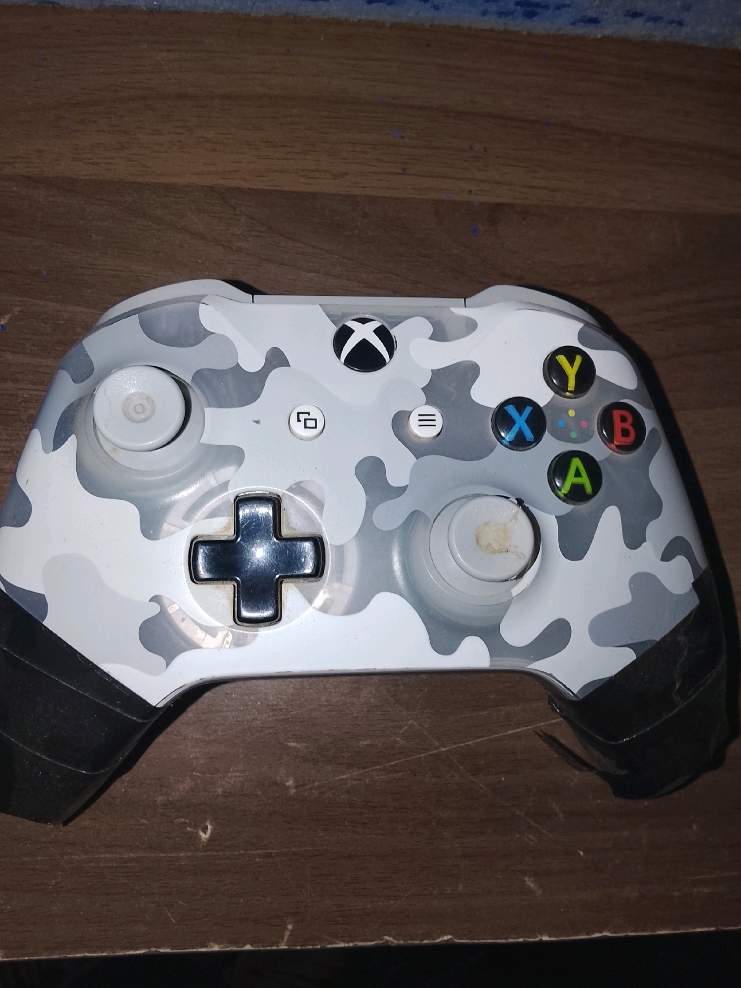 The image shows a white Xbox controller with a camouflage pattern of gray and black on a wooden surface. The controller is positioned horizontally, with the top of the controller facing the viewer. The buttons are visible, including the directional pad, analog sticks, A, B, X, and Y buttons, and the Xbox button. The grips of the controller are black. The wooden surface appears to be a table or floor, with visible grain and some scuff marks. There is no indication of any specific location within Ongata Rongai ward, Kenya, or any people present in the image.