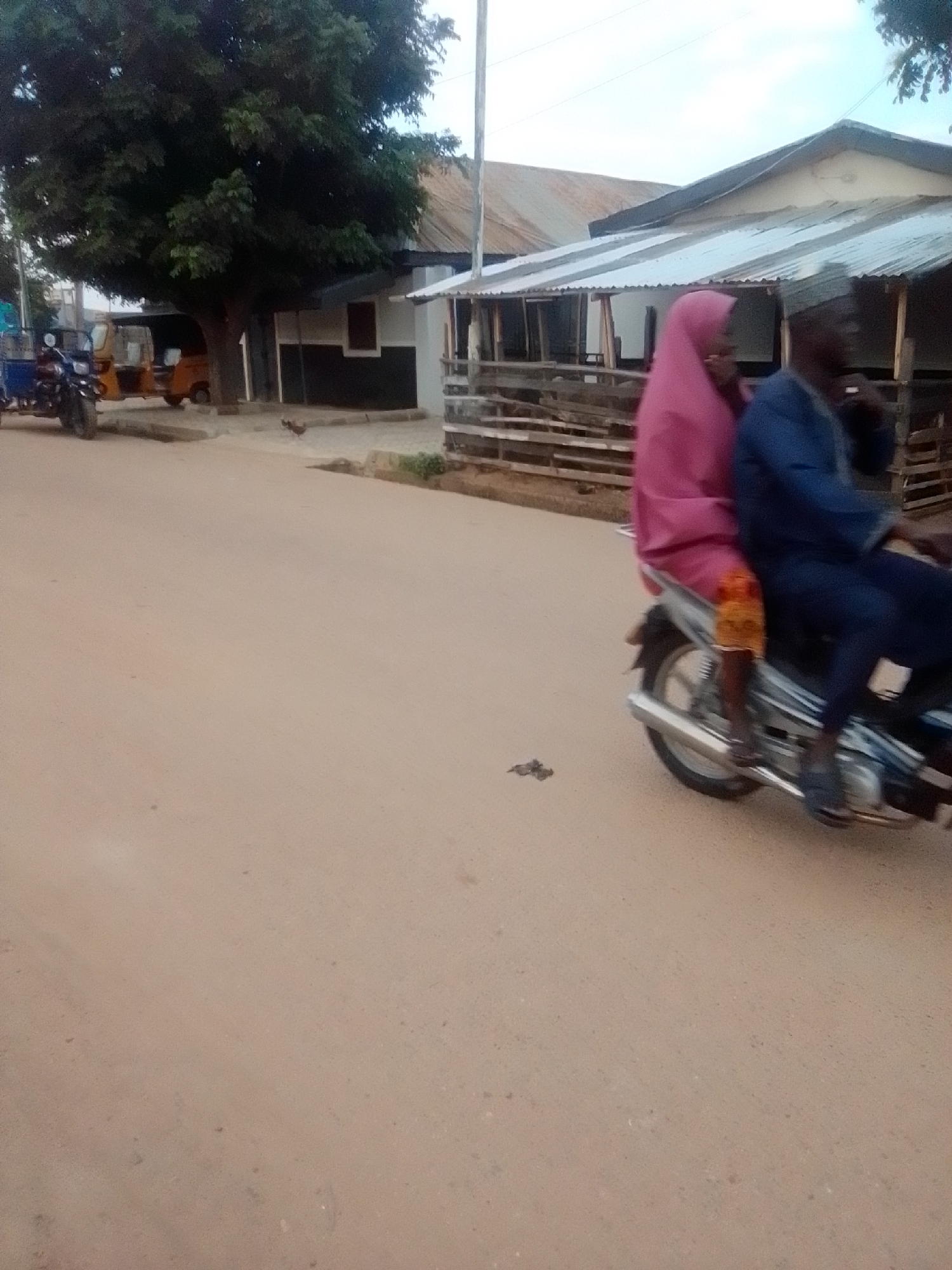 In Gombe, Nigeria, an outdoor scene depicts a street with buildings and vehicles. Two individuals are riding on a motorcycle. The person in the rear wears a pink headscarf and a patterned skirt. The person in front wears a blue jacket and dark pants. The motorcycle is silver with a black seat. In the background, there are several structures, including a building with a corrugated metal roof and a smaller building with a dark lower section and white upper section. To the left, there is a large tree with dense foliage. A yellow auto-rickshaw is parked near the buildings, and a blue motorcycle is also visible. The street is unpaved and sandy. The sky is overcast.