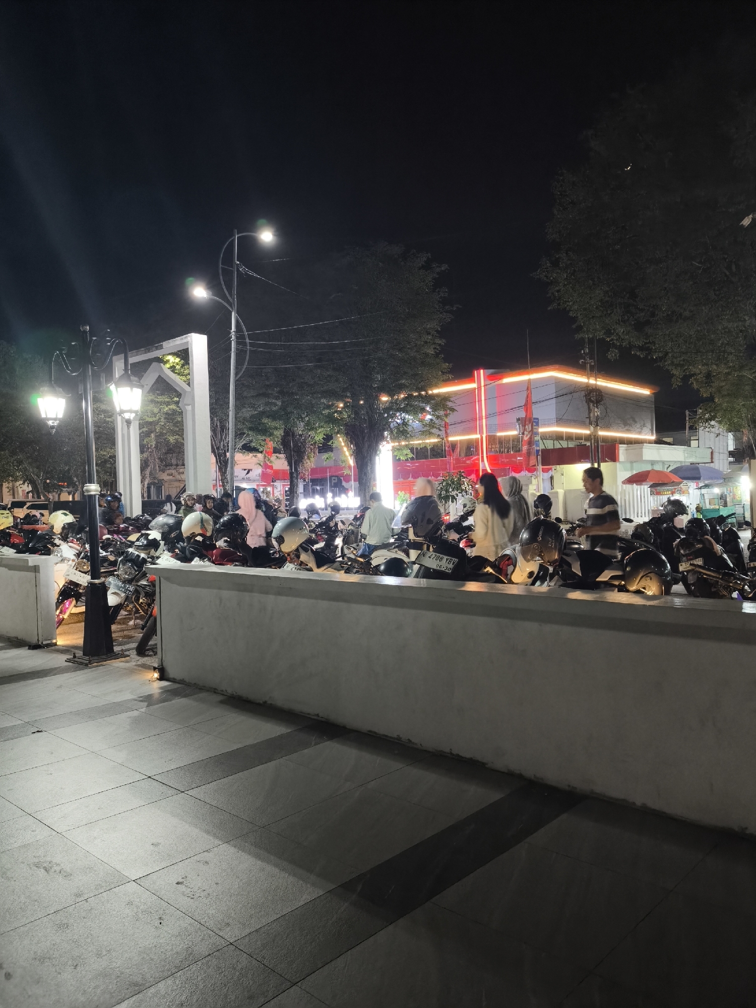 This nighttime outdoor scene captures a view of motorcycles parked in what appears to be a public area in Lumajang, Indonesia. The foreground features a tiled walkway with a white concrete barrier. Behind the barrier, a large number of motorcycles are visible, many with helmets placed on them or secured to the handlebars. Several individuals are standing near or interacting with the motorcycles, some appearing to be preparing to leave or having just arrived.
In the background, trees line the street, and a brightly lit building with orange and red neon accents stands out against the dark sky. Streetlights illuminate the area, casting long shadows. The overall atmosphere suggests a busy evening gathering or transit point in the city.