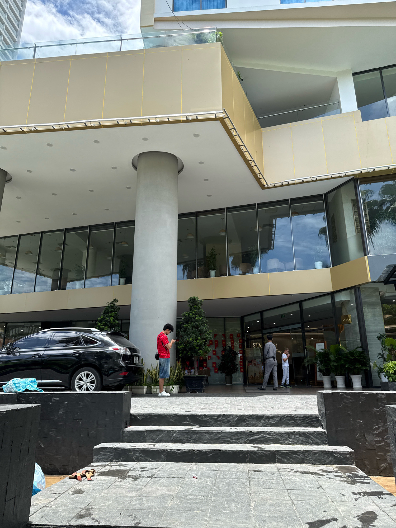 The image shows the entrance of a building in Nha Trang, Vietnam. In the foreground, there are dark gray stone steps leading up to a covered walkway. To the left of the steps, a black SUV is parked, and a blue plastic bag is on the ground. A person in a red shirt and blue shorts is standing near a large gray column, looking at their phone. Several small green trees are planted along the walkway. The building has large glass windows and a beige exterior. Further back, two people are standing near the entrance of the building. One is wearing a gray uniform, and the other is a child in a white outfit. The sky is cloudy.