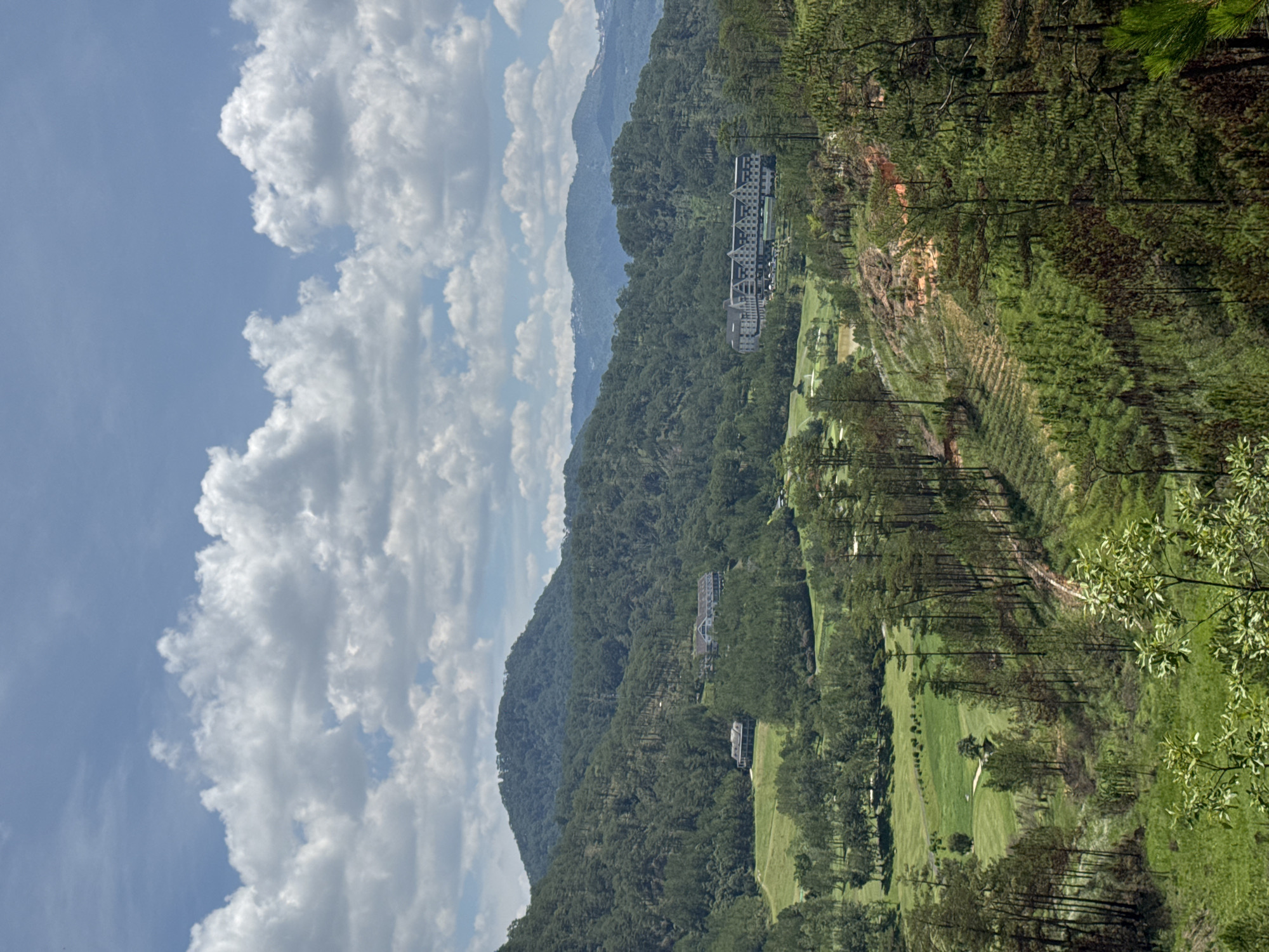 The image depicts a scenic view in Hiep Thanh, Vietnam, featuring a landscape comprised of mountains, lush green vegetation, and a cloudy sky. Several structures, possibly resorts or hotels, are nestled into the mountainside. A golf course is visible, carved into the terrain. The foreground and middle ground consist of dense forests. The sky is filled with fluffy white clouds.