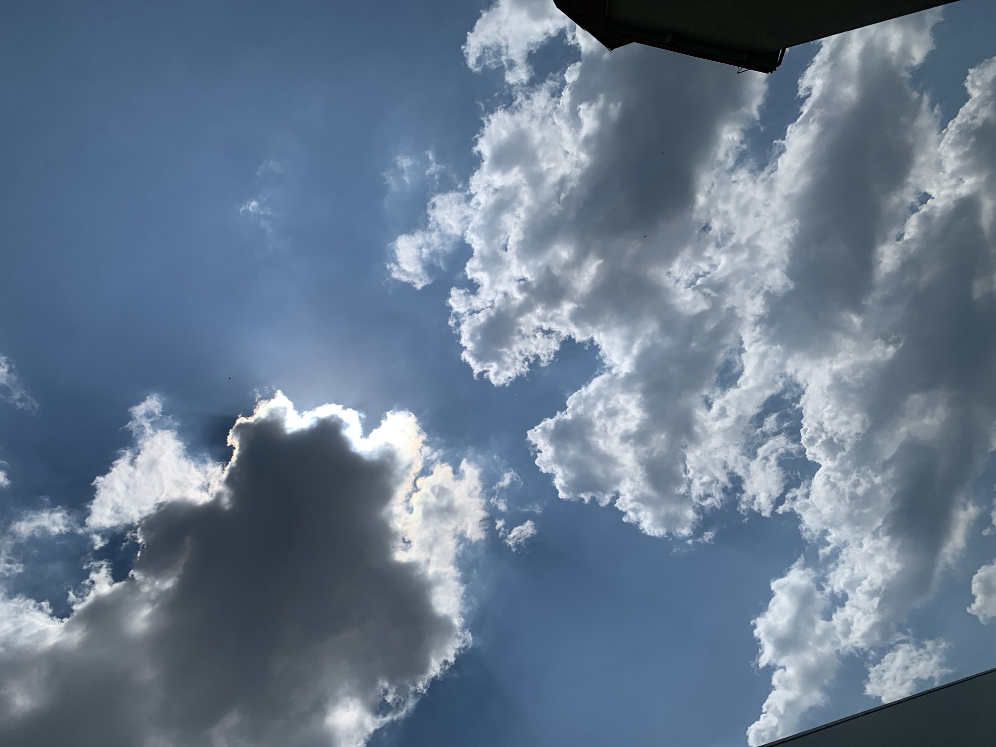 A daytime, wide-angle shot looks up at a bright blue sky filled with a mix of dark gray and white cumulus clouds. The sun is partially obscured by a large, dark cloud in the upper left quadrant, creating a bright halo effect and rays of light that penetrate the surrounding sky. Several smaller, white clouds are scattered throughout the scene. In the lower right corner, the top of a building with a dark, sloped roof is visible, along with a sliver of a lighter colored wall. In the far lower left corner, a portion of a light-colored building or structure is visible. Two small dark specks, possibly birds, are present in the sky. The scene is a street view in Kolomyia, Ukraine.