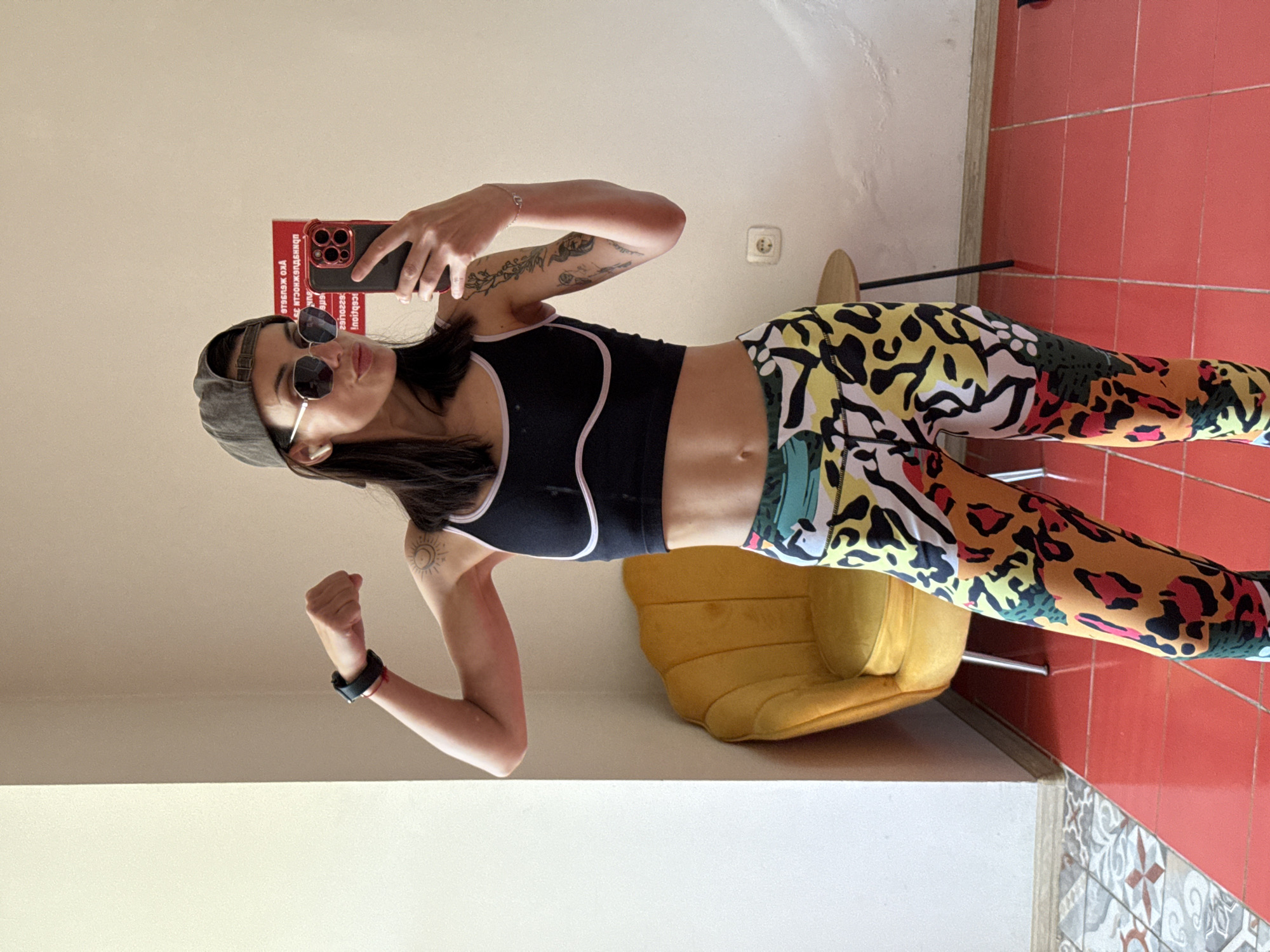 A woman is taking a selfie in a room with a red tiled floor and a yellow chair. She is wearing sunglasses, a grey baseball cap, a black sports bra with white trim, and brightly patterned leggings. She is holding a red cell phone in her right hand and flexing her left arm, which has a black wristband. Tattoos are visible on both arms. A white wall is visible in the background. An electrical outlet is visible on the wall. Based on the book "ОБИЧАЙ СЕ ПОД ВОЛЯТА И АЗ ЩЕ ТЕ НАМЕРЯ", this image may have been taken in the city of Pleven, Bulgaria.