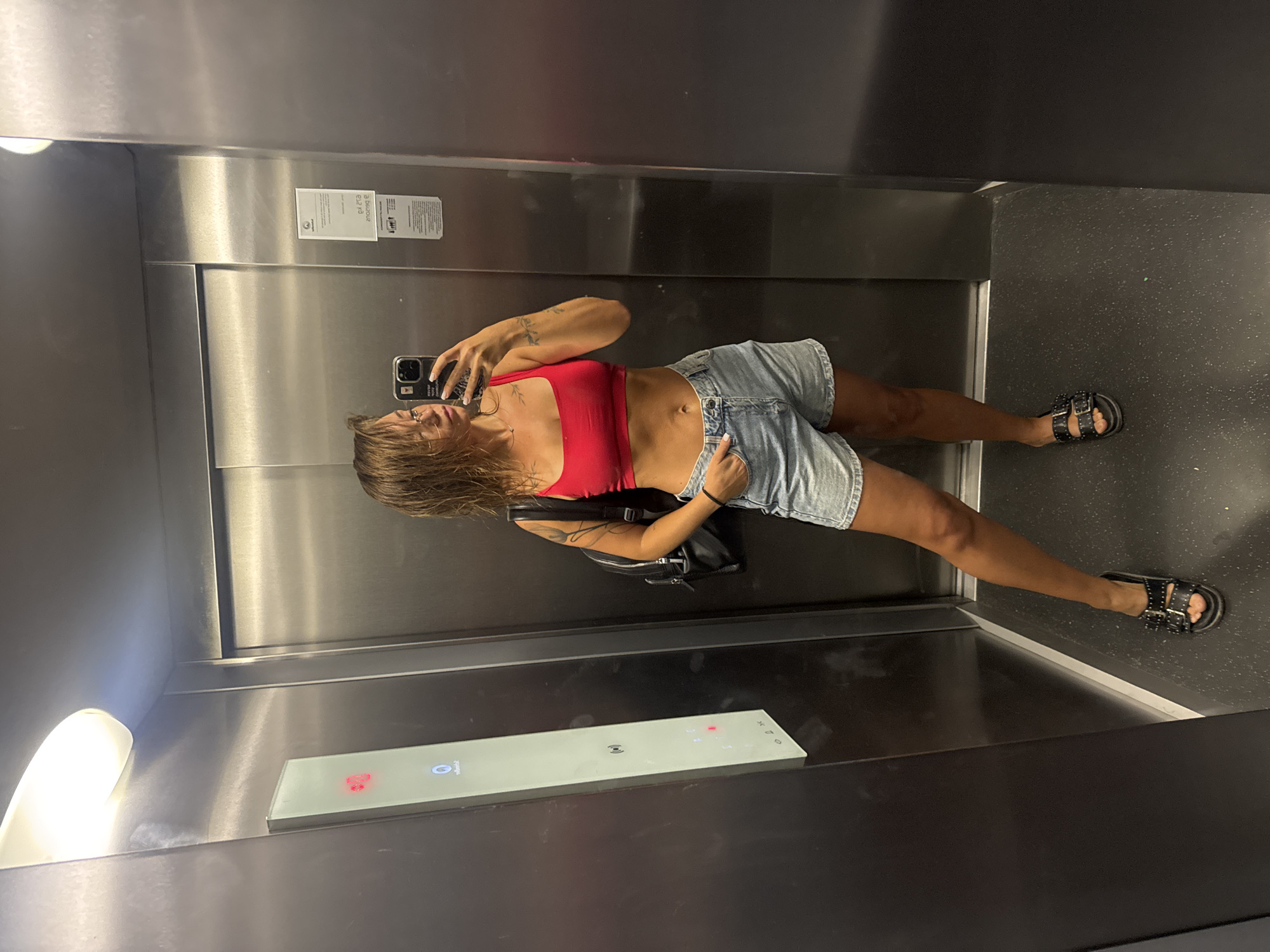 A young woman takes a mirror selfie in an elevator. She is wearing a red crop top and denim shorts, with a black backpack slung over her shoulder. She has wet hair and is wearing black sandals. The elevator has stainless steel walls and a lighted panel with buttons. The woman is standing in the center of the elevator, facing the mirror. The scene appears to be in Budva, Montenegro, given the context provided in the prompt, although there are no visual cues in the image to confirm this location.