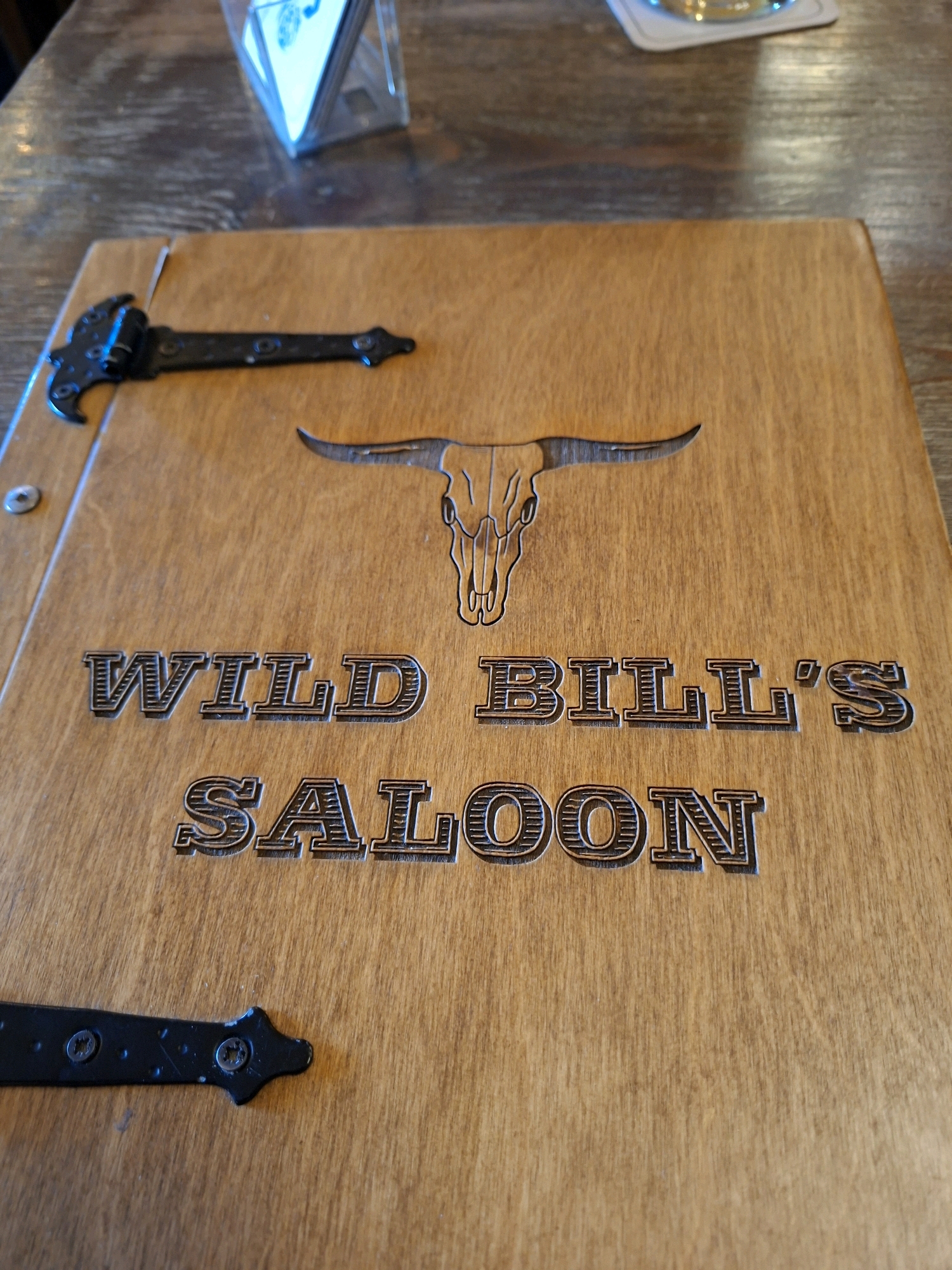 A close-up shot shows the wooden cover of a menu at "Wild Bill's Saloon." The cover features an embossed image of a longhorn skull above the embossed text "WILD BILL'S SALOON." Black metal hinges are attached to the left side and bottom of the menu cover. The menu is resting on a wooden table. In the upper background, a clear plastic holder contains several white cards, one of which has a blue design. The location, "Wild Bill's Saloon" in Rastatt, Germany, is not explicitly visible in the image itself.