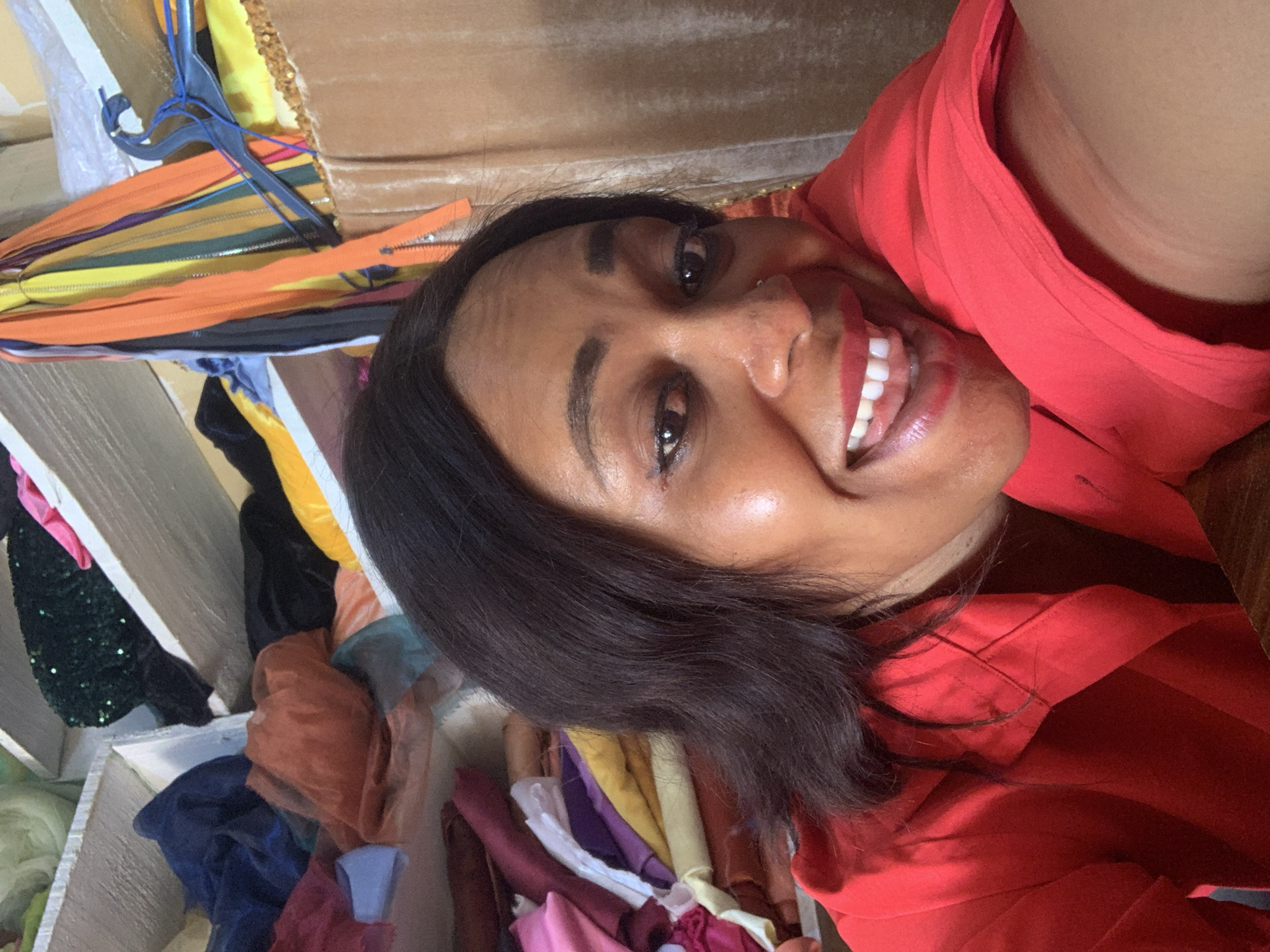 In the city of Sabon Tasha, Nigeria, a Black woman with dark hair is captured in a close-up, smiling selfie. She is wearing a vibrant red collared shirt, and her makeup includes bright red lipstick and eyeliner. The background is a cluttered shelf filled with various fabrics, including navy blue, orange, pink, and yellow materials, along with a collection of colorful zippers. The setting appears to be a workshop or storage area for textiles.