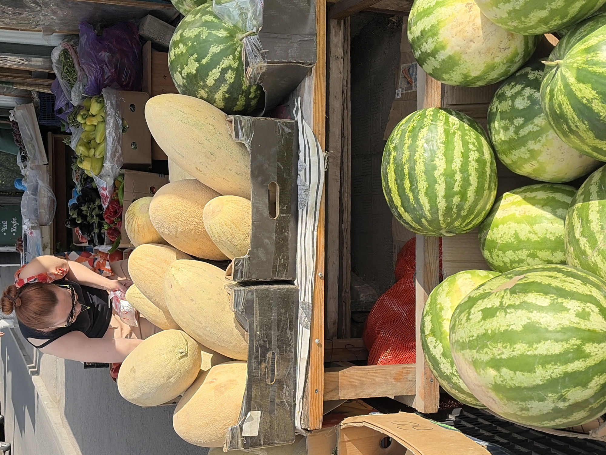 A woman with auburn hair pulled into a bun, wearing sunglasses and a black tank top with a colorful scarf around her neck, is bent over a display of large, pale yellow melons at an outdoor market. She is looking down at the melons, and her hands are near a white plastic bag. Behind her, a table is laden with more produce, including peppers, eggplants, and other fruits and vegetables. In the foreground, a large pile of watermelons with their characteristic green stripes is visible. The scene appears to be a sunny day, with shadows cast by the stall and surrounding structures.