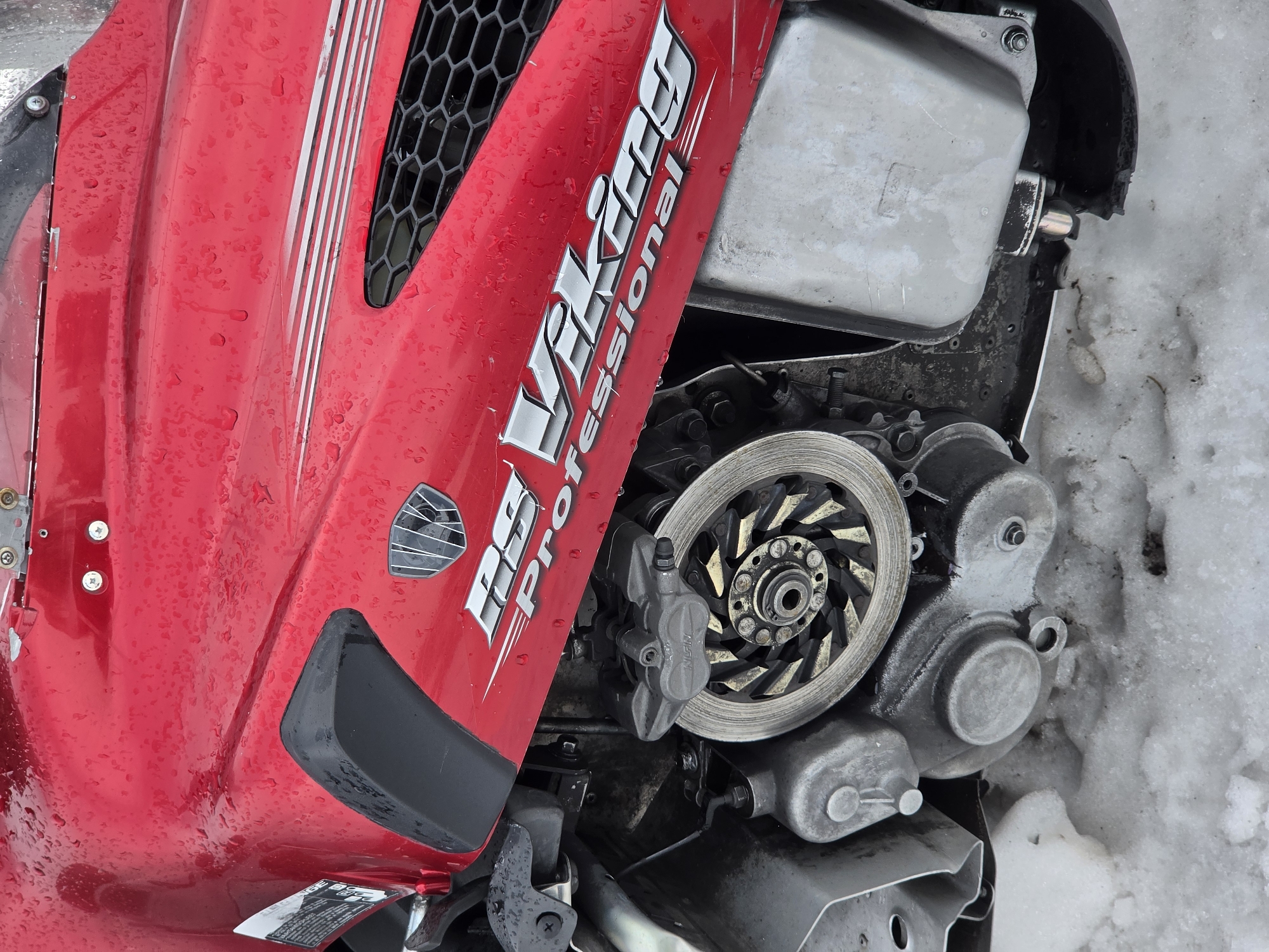 This is a close-up, low-angle, eye-level shot of the side of a red snowmobile, partially covered in snow, in Terskol, Russia. The snowmobile's body is wet, with visible water droplets, and it has a "RS Viking Professional" decal on the side. A portion of the engine, including a disc brake and caliper, is visible. The background is blurred and suggests an outdoor environment with snow. There are no people or animals in the image. The focus is on the mechanical and exterior details of the snowmobile.