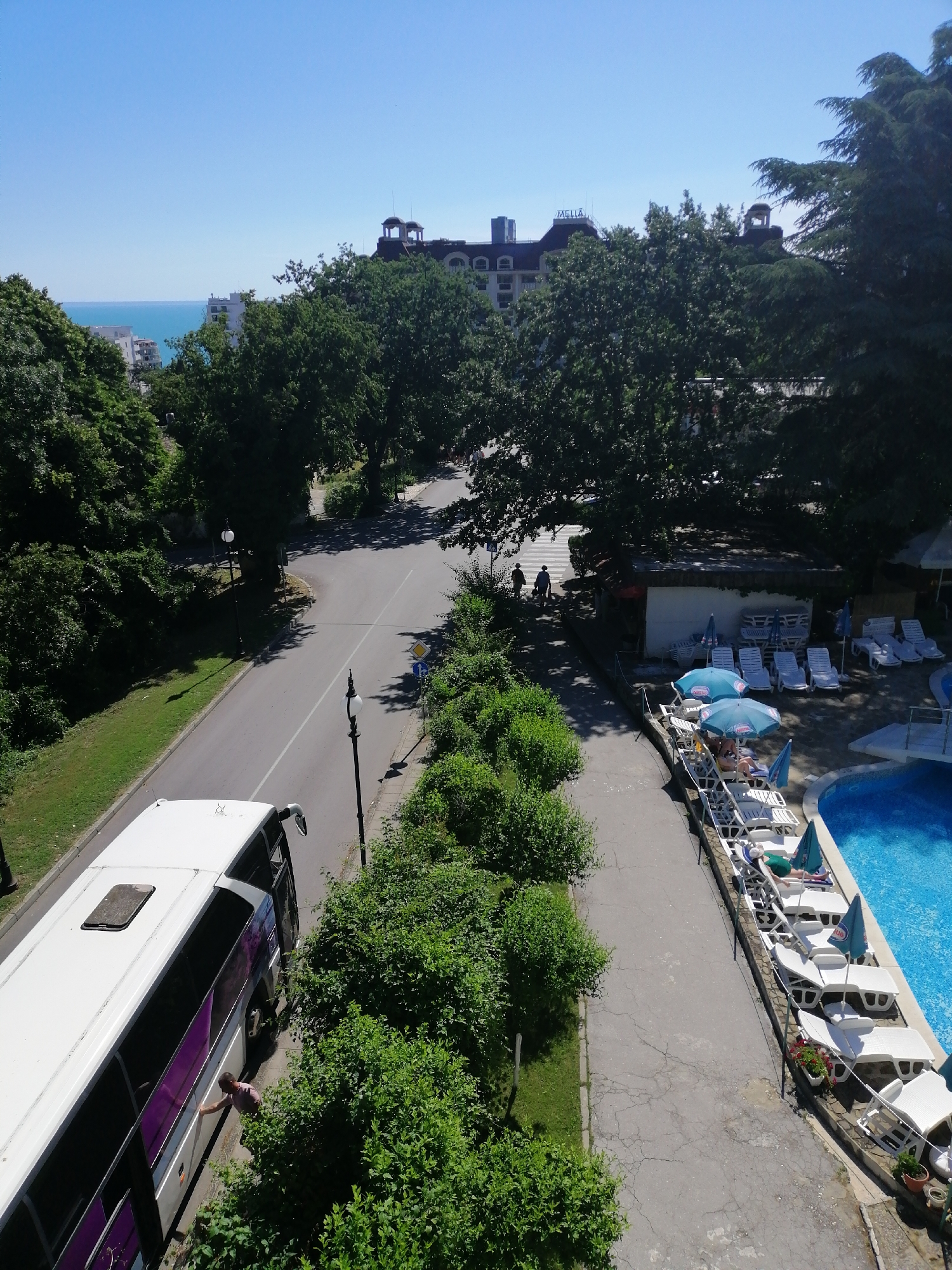 In Varna, Bulgaria, a white bus with purple accents is parked on the side of a road lined with trees. A man is visible near the bus, reaching towards its side. On the opposite side of the road, a swimming pool area is set up with lounge chairs and umbrellas, and a few people are relaxing. In the background, a large hotel with the name "MELIA" visible on its roof is situated, with the sea beyond it. The sky is clear and blue.