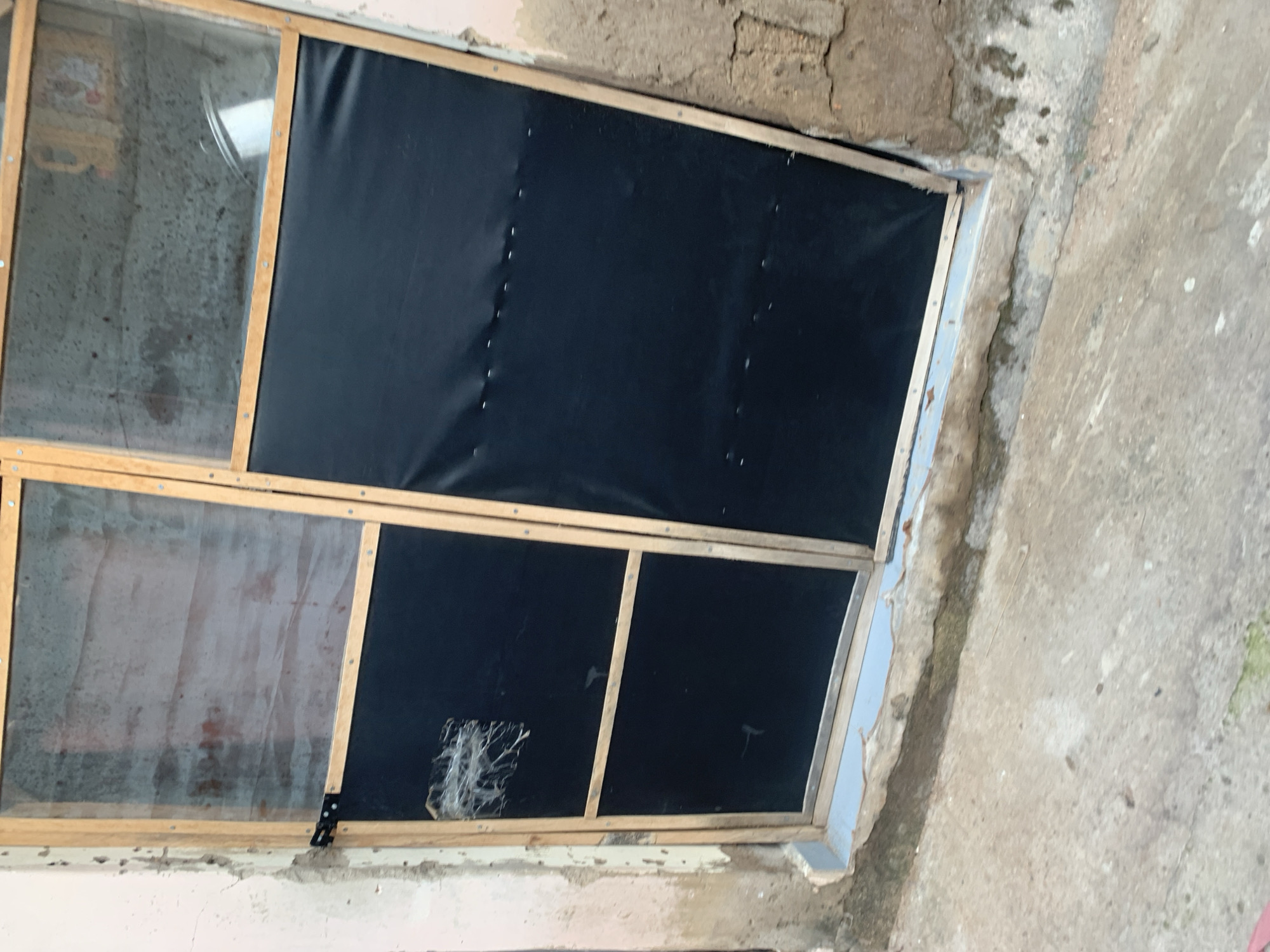 The image shows a weathered wooden framed window or door with a black tarp covering most of it. The left side of the frame has a small section with a torn screen, and above that, a dusty glass pane. The window is set into a rough, light pink wall on the left and a darker, textured wall on the right. The ground in front of the window is concrete, showing cracks and some green moss. The scene appears to be an exterior shot, possibly in a less maintained area of Jalingo, Nigeria, given the rustic construction and evident wear and tear.