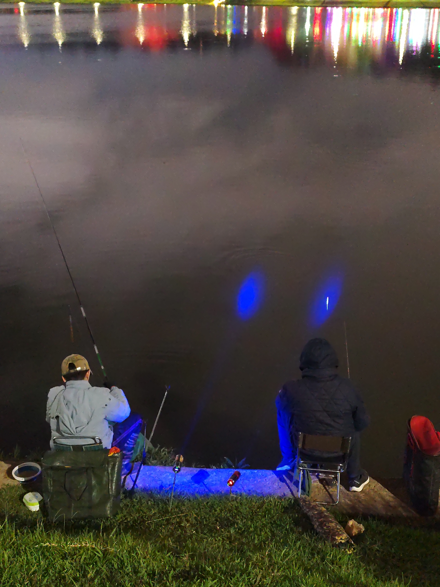 Two people are fishing at night on a body of water, likely a lake, with a shoreline illuminated by blue lights. The reflections of colorful lights from the city are visible on the water in the background. The person on the left is wearing a light-colored jacket and a hat, and is seated on a folding chair. To their left is a green fishing bag and other fishing-related items. The person on the right is wearing a dark hooded jacket and is seated on a metal folding chair. Red boots or a bag are visible to their right. Two blue lights are visible in the water in front of the person on the right. The scene is situated in Da Lat, Vietnam, identified by the specific arrangement of lights and the overall atmosphere.