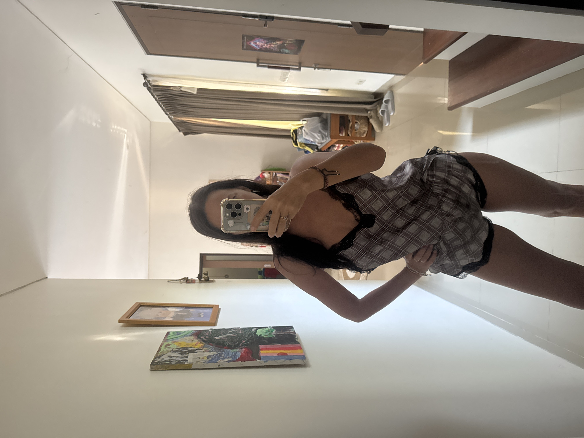 A woman is positioned in front of a large mirror, taking a photograph of herself using a smartphone. She is wearing a plaid, two-piece outfit with black lace trim. The room includes various decor items on the wall, such as paintings and framed pictures. There is a door at the end of the hallway, along with items like shoes and a bag placed on a shelf or table. The setting is an interior space, possibly a residence or apartment. The location in the city is Unknown, Unknown.