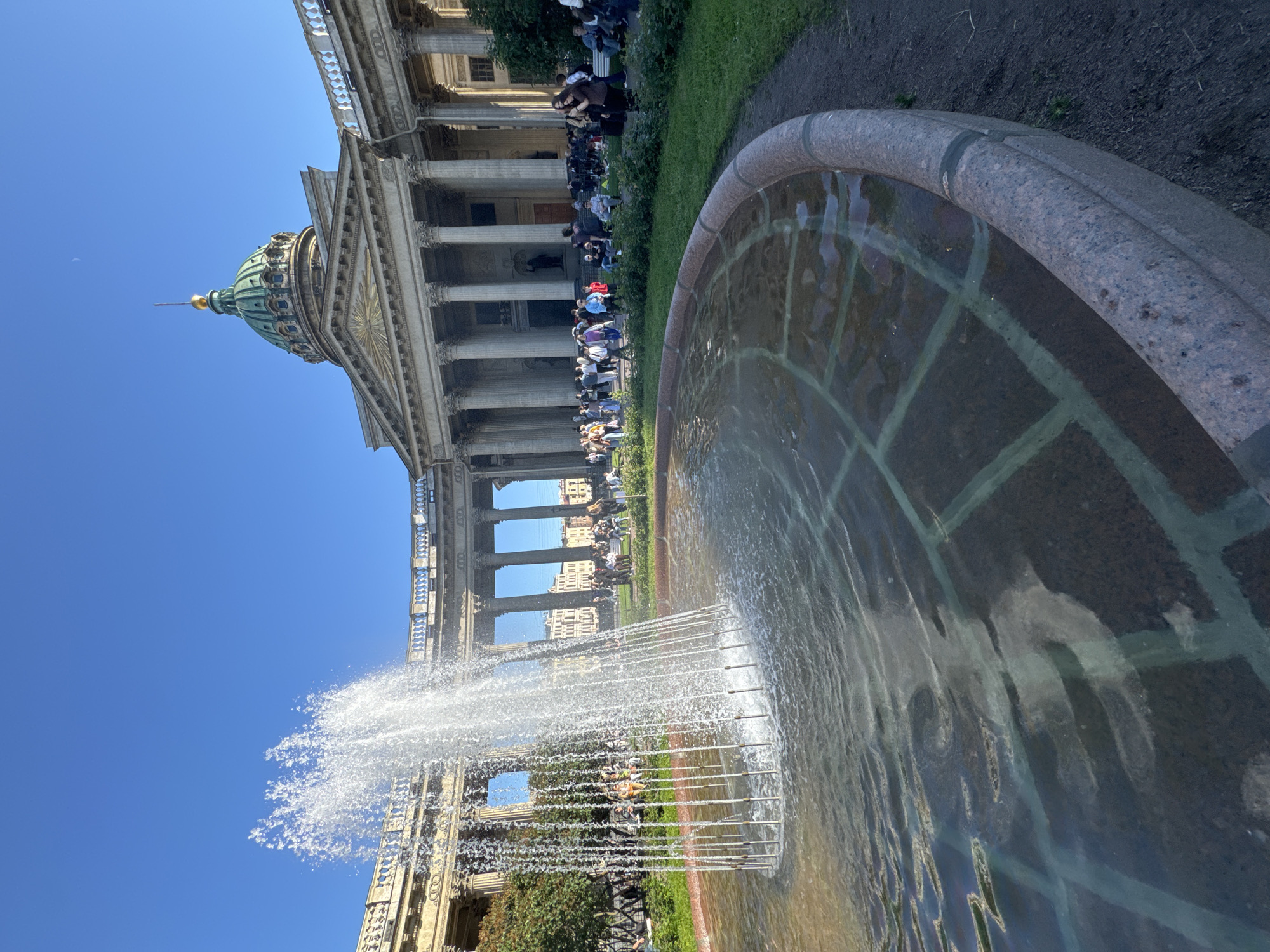 This image captures a sunny day in Saint Petersburg, Russia, likely during the daytime given the bright sunlight. The foreground features a large, ornate fountain, with water spraying upwards in a delicate mist. The fountain basin is made of a textured, reddish-brown stone with a patterned interior.
Behind the fountain, a grand classical building dominates the scene. Its facade is adorned with numerous tall columns and intricate architectural details. A prominent green dome, topped with a golden spire, rises from the building, suggesting it is a significant religious or historical structure, possibly the Kazan Cathedral. A line of people are visible seated or standing along the grassy area in front of the building, perhaps tourists or locals enjoying the public space. The sky is a clear, vibrant blue, indicating fair weather.
In the background, other buildings can be seen, hinting at the urban environment of Saint Petersburg. The overall impression is one of a peaceful, culturally rich urban setting on a pleasant day. The image is oriented sideways, with the ground level appearing on the left and the sky on the right.