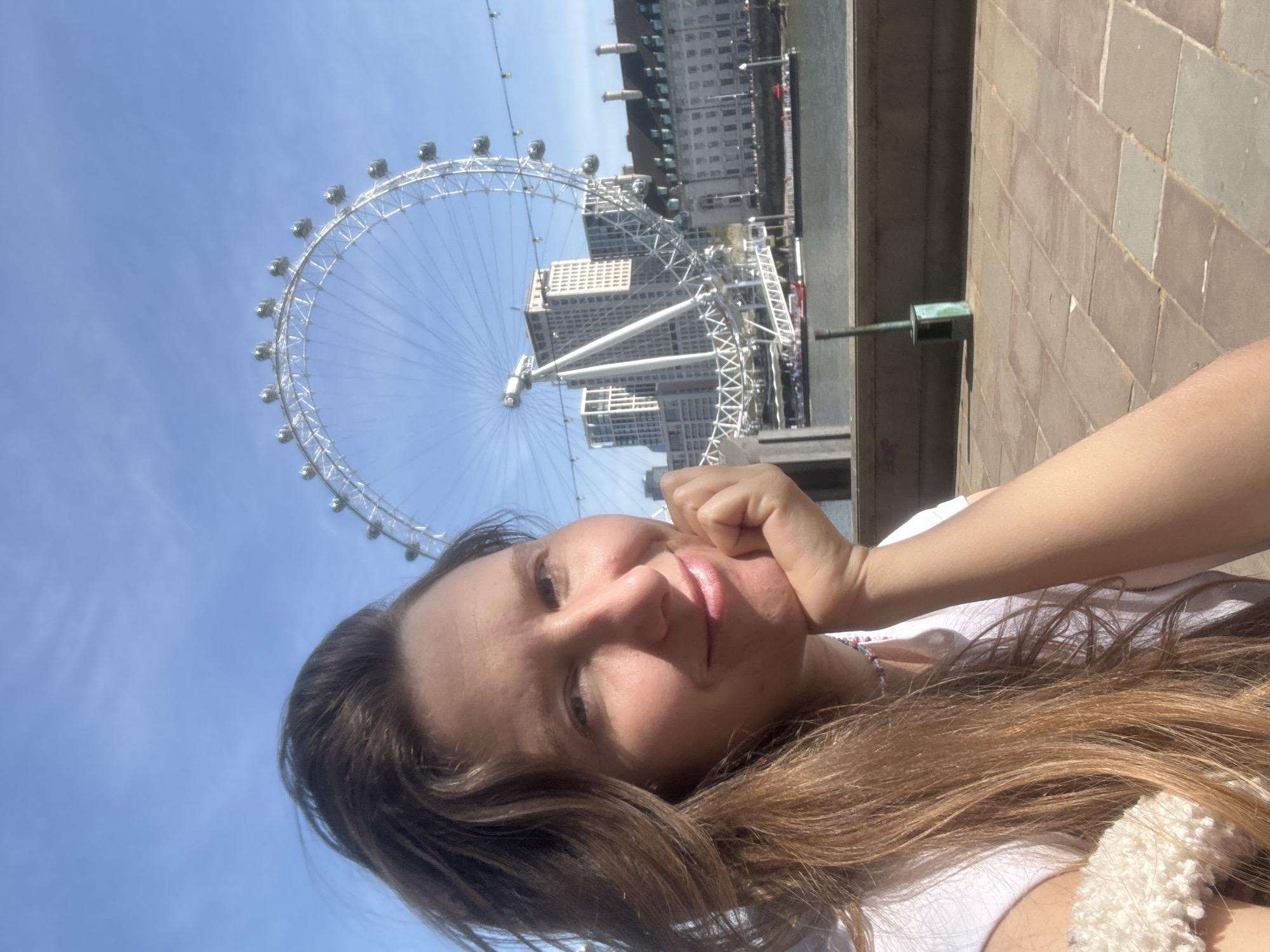 A selfie featuring a woman with brown hair and a white shirt, with the London Eye Ferris wheel in the background. The image captures a section of a building behind the Ferris wheel, as well as the edge of a brown-tiled pathway or structure in the foreground. The location is London, United Kingdom.