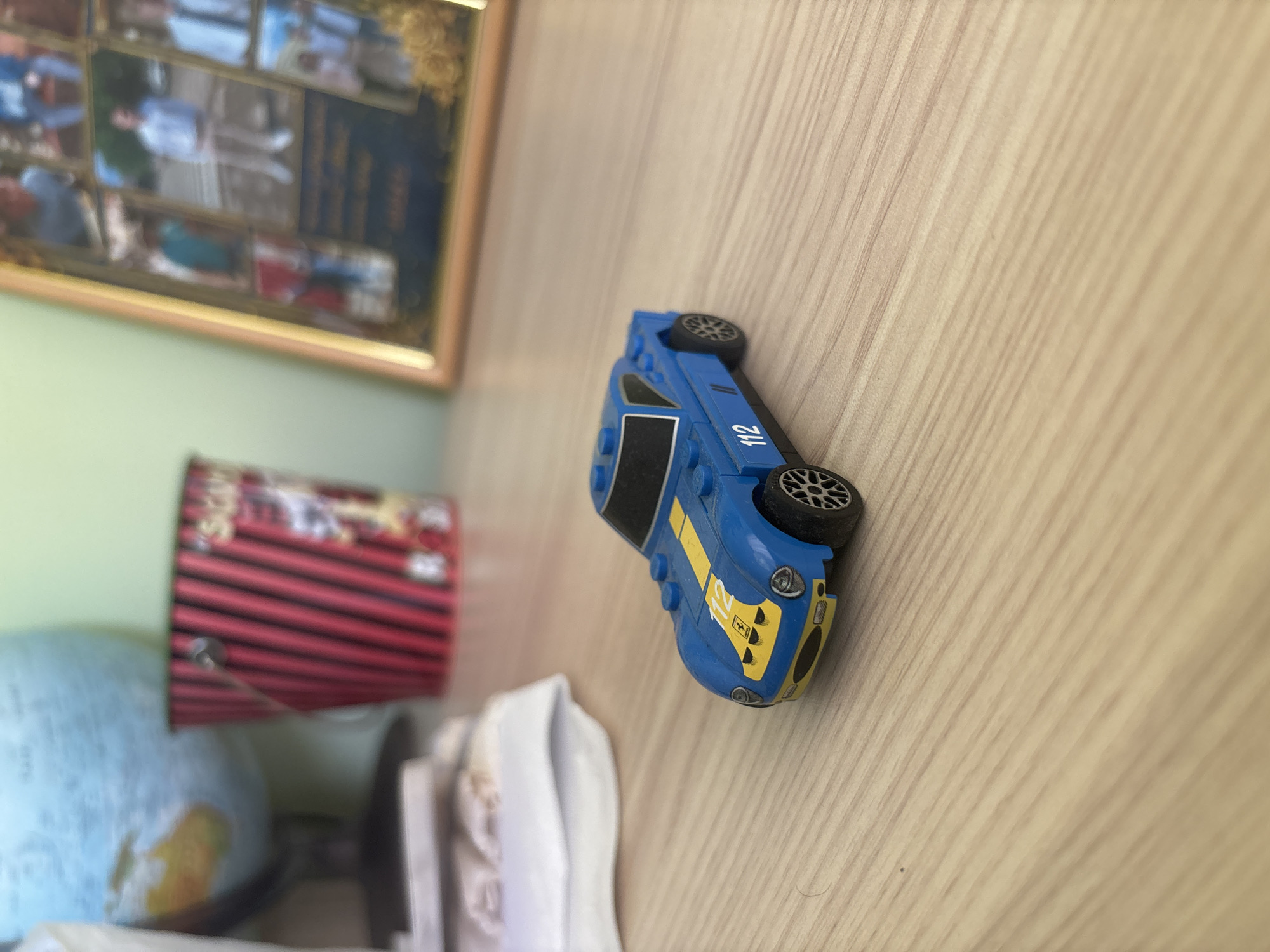 The image presents a tilted, indoor scene from Blagoevgrad, Bulgaria, featuring various objects arranged on a light-colored wooden surface with visible grain lines, likely a desk or table. The central focus is a blue toy racing car, resembling a Lego or similar building block vehicle, positioned prominently. This car has yellow accents, including a stripe on its side with the number "112" clearly printed in white. A yellow shield featuring a black prancing horse, indicative of the Ferrari logo, is visible on its front hood. The car is equipped with black tires and silver-colored hubs.
In the background, on the left, a framed picture collage is mounted on a light green wall. The individual photographs within the frame are blurry but appear to be personal or family snapshots. Below the framed picture, a red and black striped cylindrical object, possibly a pencil holder or mug, is partially visible. Further towards the bottom left, a light blue globe with yellow and brown landmasses is partially seen. Some crumpled white fabric or paper is visible in the lower central part of the frame. The lighting suggests it is daytime. The overall perspective is a downward shot, rotated, making the wooden surface appear as a vertical plane.