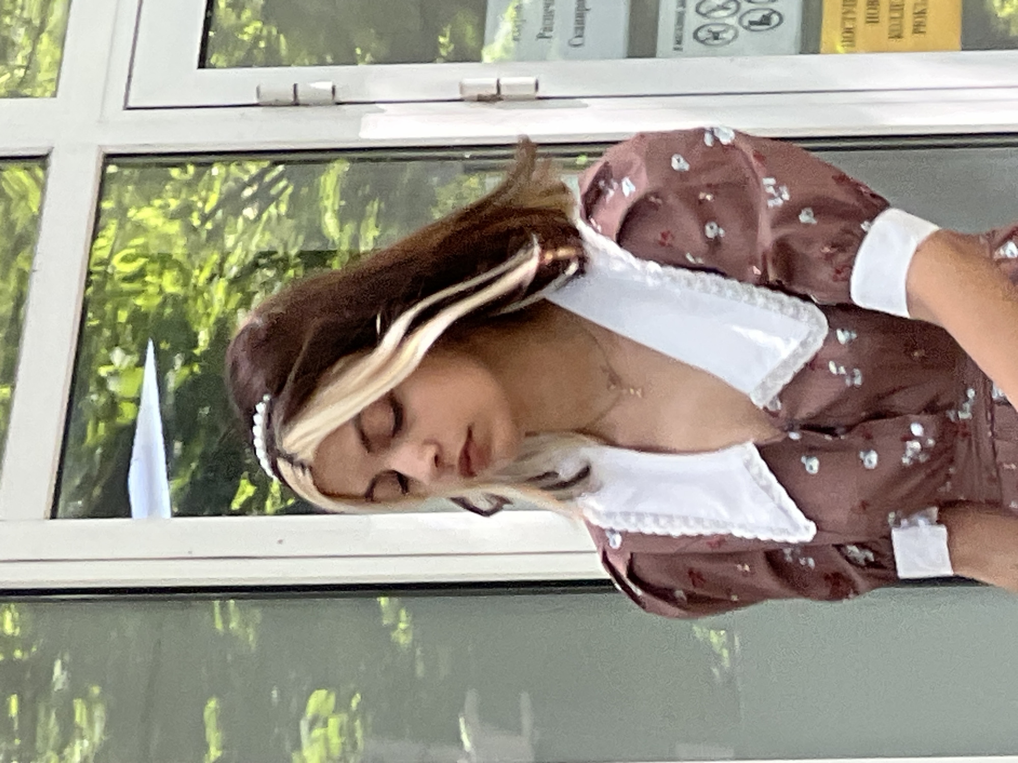 A young person with dark hair streaked with blonde is captured in a medium shot, looking downwards with their eyes closed. They are wearing a brown floral-print dress with a large white collar and white cuffs. A pearl headband adorns their hair. The background consists of a glass building with reflections of green trees, suggesting an outdoor setting in a city. The overall impression is one of a casual, daytime scene. This image was taken in Krasnodar, Russia.