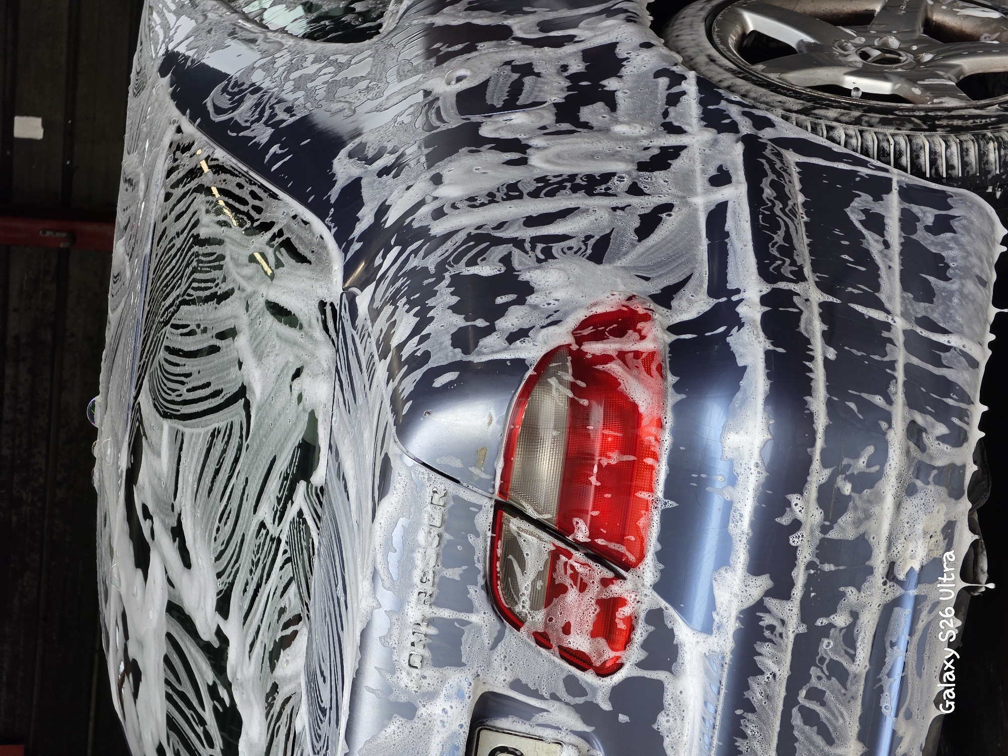 A close-up, low-angle shot captures the rear quarter panel of a dark blue car being washed. The car is covered in white soap suds, with streaks and patterns formed as the water runs down its surfaces. A red taillight is visible on the left side of the frame, partially obscured by the suds. The car's badge, "CHRYSLER," is subtly visible on the trunk. A portion of a silver alloy wheel and tire is visible on the right. The background is dark and indistinct, suggesting an indoor washing area or shaded outdoor location. The bottom left corner of the image contains a watermark reading "Galaxy S26 Ultra." The scene is devoid of any people or specific urban landmarks to pinpoint a location within Plovdiv, Bulgaria.