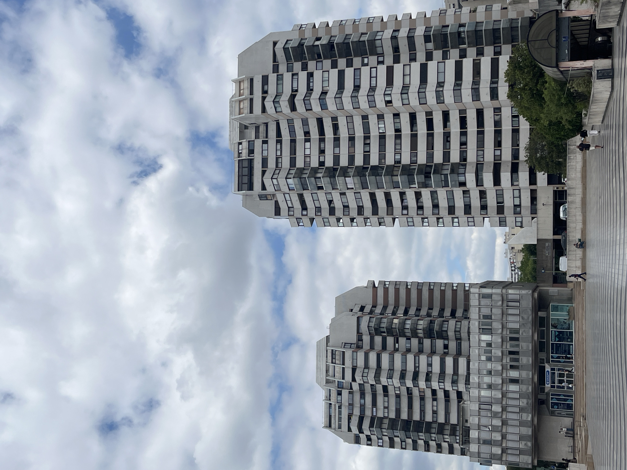 This image shows a view of two modern, angular apartment buildings in Créteil, France. The buildings are tall and made of concrete and glass, with a repeating pattern of windows that give them a textured appearance. The building on the right is closer to the foreground and appears to be part of a complex with a lower section that has an entrance and a curved roof structure. A few people are visible walking on the paved area in front of the buildings, some are also visible in the background. One person is walking to the right on the pavement, and another is sitting on a ledge. A white van is parked near the entrance of the building on the right. The sky is filled with scattered clouds, suggesting it is daytime, possibly late morning or afternoon. The lighting is diffused, indicating it might be partly cloudy. The perspective is from a low angle looking up at the buildings, emphasizing their height and imposing architecture. There are no discernible emotions conveyed in the image, which focuses on the architectural elements and the urban setting.