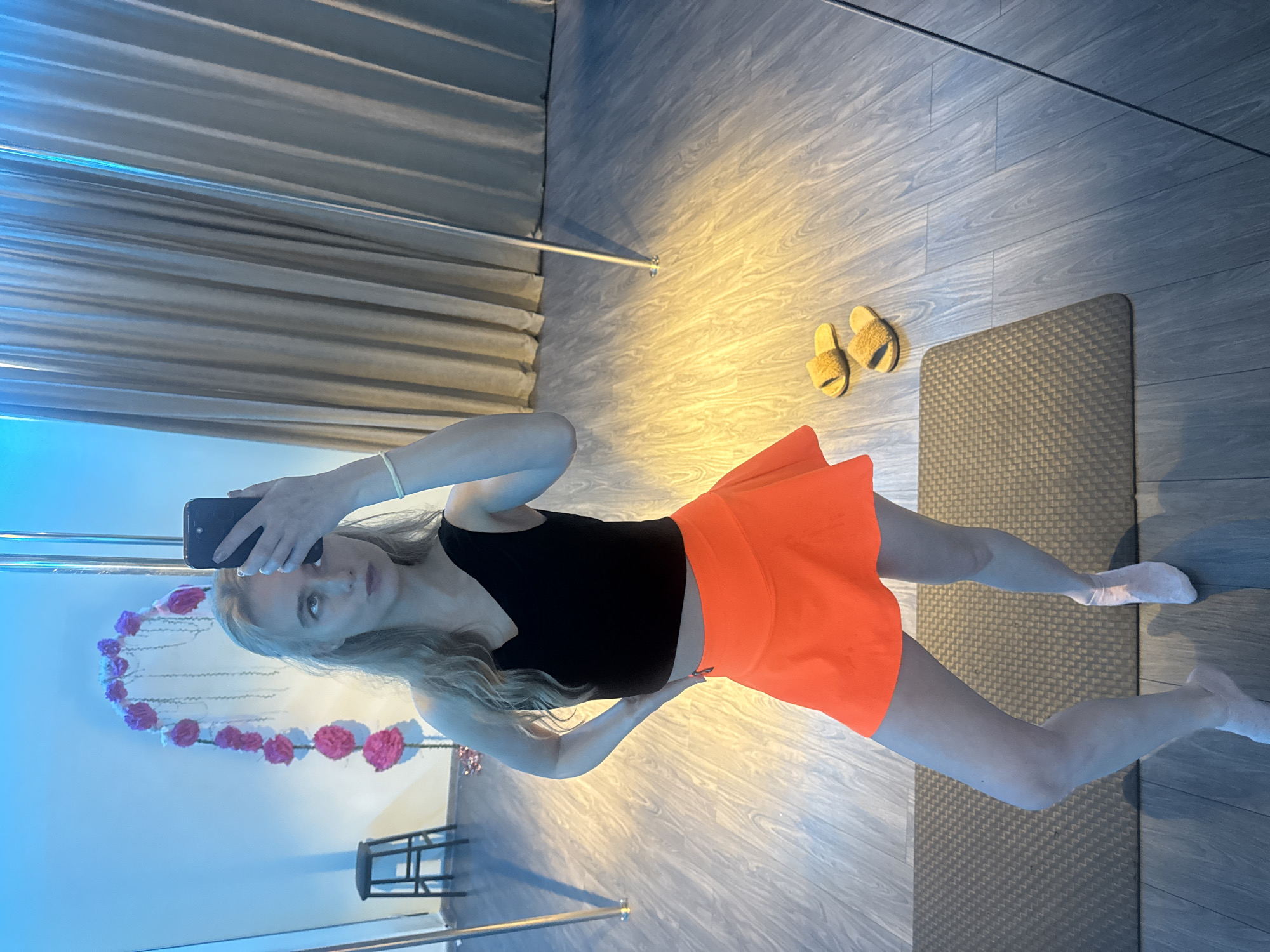A young woman is standing on a gray wood-look floor, facing a mirror, and taking a selfie with a black smartphone. She is wearing a black tank top and a bright orange skirt. She is barefoot and standing on a gray mat.
Behind her, the mirror reflects a metal pole with a heart-shaped decoration of pink flowers, a small black stool, and a blue wall. To the right of the mat, there are a pair of yellow slippers on the floor.
The background shows gray, wavy curtains and a wall with wood-look paneling. The location is Unknown, Unknown.