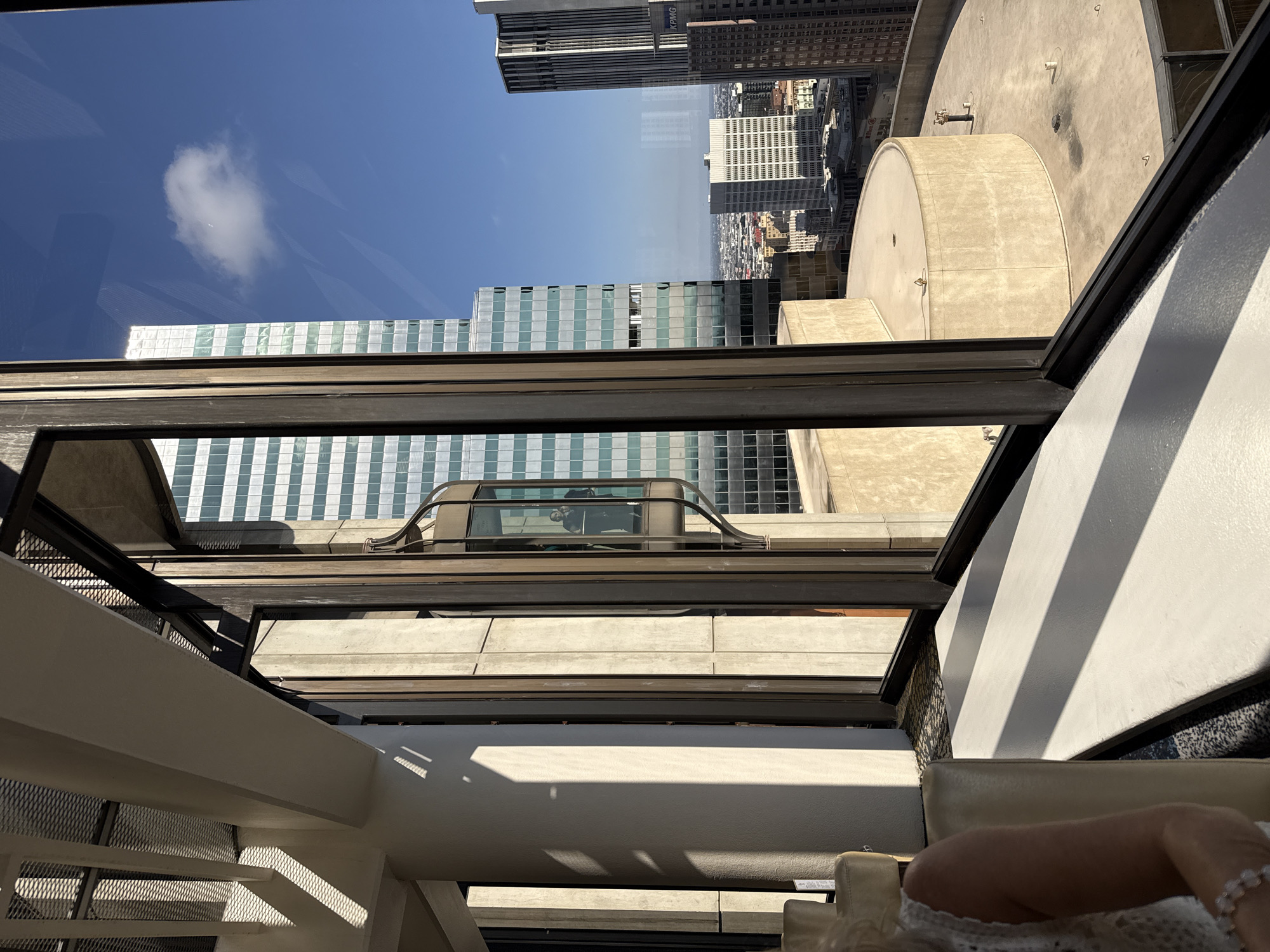 The photograph captures a view from an elevated vantage point, likely a high-rise building, looking out over a cityscape. In the foreground, to the left, a portion of a person's shoulder and arm is visible, draped in white fabric, possibly lace. They are seated on a pale-colored chair or sofa. The interior architectural elements, such as a white beam with a textured underside and a concrete column, cast sharp shadows due to the bright sunlight.
A prominent feature is a glass elevator, visible through large, framed windows, with at least two individuals inside. The elevator is sleek and modern, designed to showcase the panoramic views. Beyond the immediate interior, the exterior cityscape unfolds. Several tall buildings dominate the view, characterized by their reflective glass facades and grid-like patterns. One building, in particular, has a distinctive turquoise-tinted glass. The sky is a clear, bright blue, with a single, fluffy white cloud.
In the mid-ground, a curved, multi-tiered outdoor space with beige, concrete-like surfaces is visible, suggesting a terrace or rooftop area of a building. Further in the distance, a dense urban landscape stretches to the horizon, with a variety of buildings of different heights and architectural styles. The overall impression is one of a modern urban environment bathed in bright sunlight. The presence of the reflective skyscrapers and the general urban sprawl strongly suggests a location within the downtown area of a major city. Based on the architectural style of the buildings and the characteristic density of the urban landscape, this scene is likely located in the city of Los Angeles, United States of America.