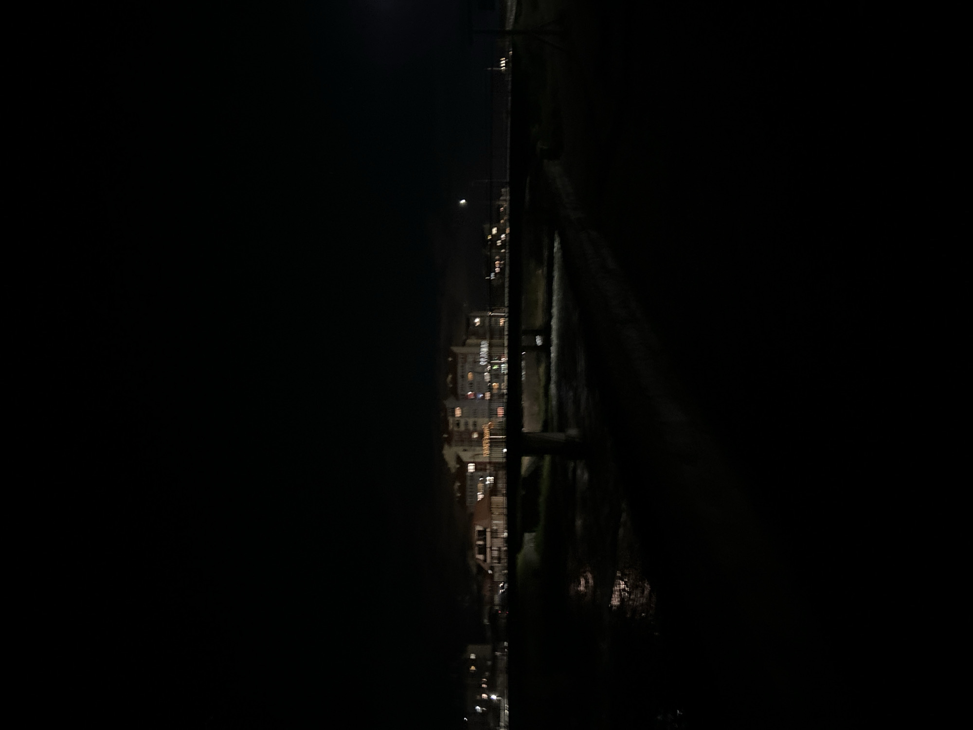 The image shows an urban night scene with lights from buildings visible. A dark body of water is present on the right side of the frame. A dark object, possibly a bridge or structure, spans across the view over the water.