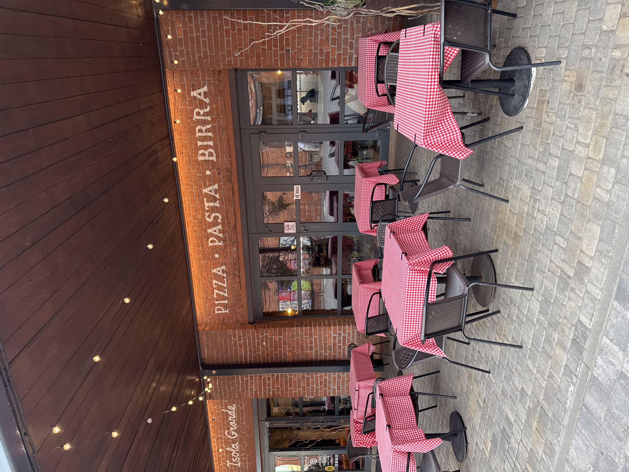 The image shows an outdoor seating area of a restaurant with red and white checkered tablecloths. Tables are set with chairs on a cobblestone patio. The restaurant's exterior is made of red brick, and a sign on the wall reads "PIZZA PASTA BIRRA" and "Isola Grande." String lights are visible hanging above the seating area. The doors to the restaurant have glass panels, and reflections of people and the interior are visible. There is text on the doors, including "ВХОД" (ENTRANCE) in Russian. The overall impression is of a pleasant, casual dining atmosphere. The lighting suggests it might be daytime, possibly overcast, or early evening with artificial lighting activated. The location is identified as Saint Petersburg, Russia.