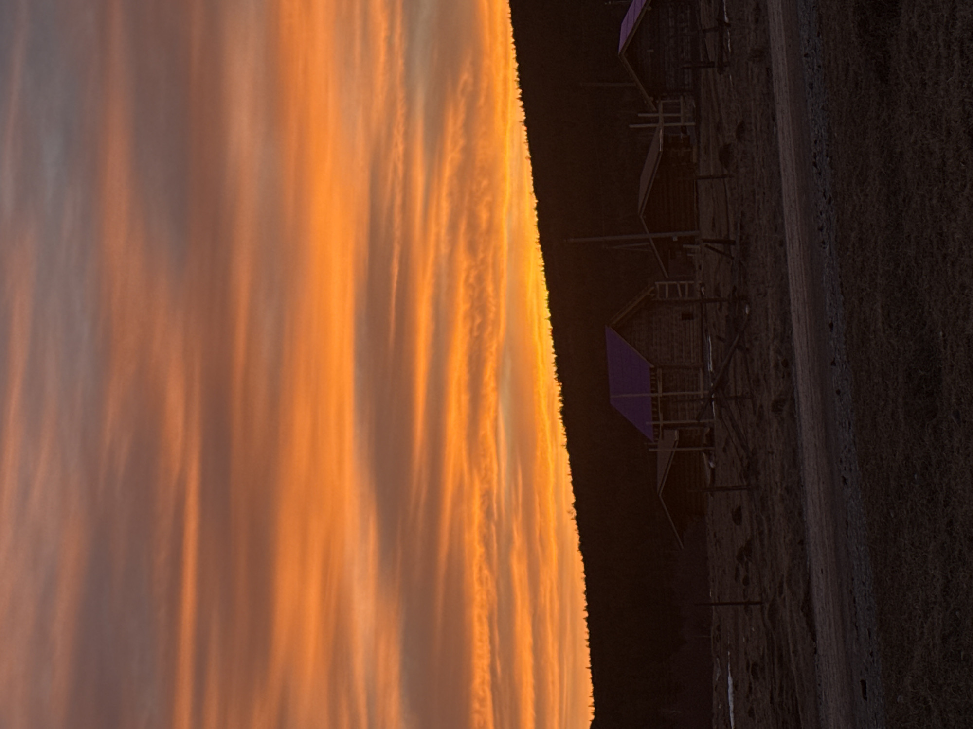 The image displays a rural landscape in Tyulyuk, Russia, during twilight hours. The upper two-thirds of the frame are dominated by a sky exhibiting strong orange and reddish-orange hues with pronounced horizontal cloud striations. Below this vibrant sky, a dark, silhouetted coniferous forest line extends horizontally across the mid-ground. In front of the forest, a small cluster of dark, wooden structures, likely cabins or houses, is visible. Two of these structures distinctively feature purple-colored roofs. Basic wooden fencing and utility poles are discernible among the buildings. The foreground consists of dark, uneven terrain, appearing sparsely vegetated and possibly frozen, with a faint unpaved road or track running horizontally across the lower portion. Scattered patches of snow or frost are evident on the ground, particularly to the left. No individuals, explicit actions, or interactions are present in the scene.