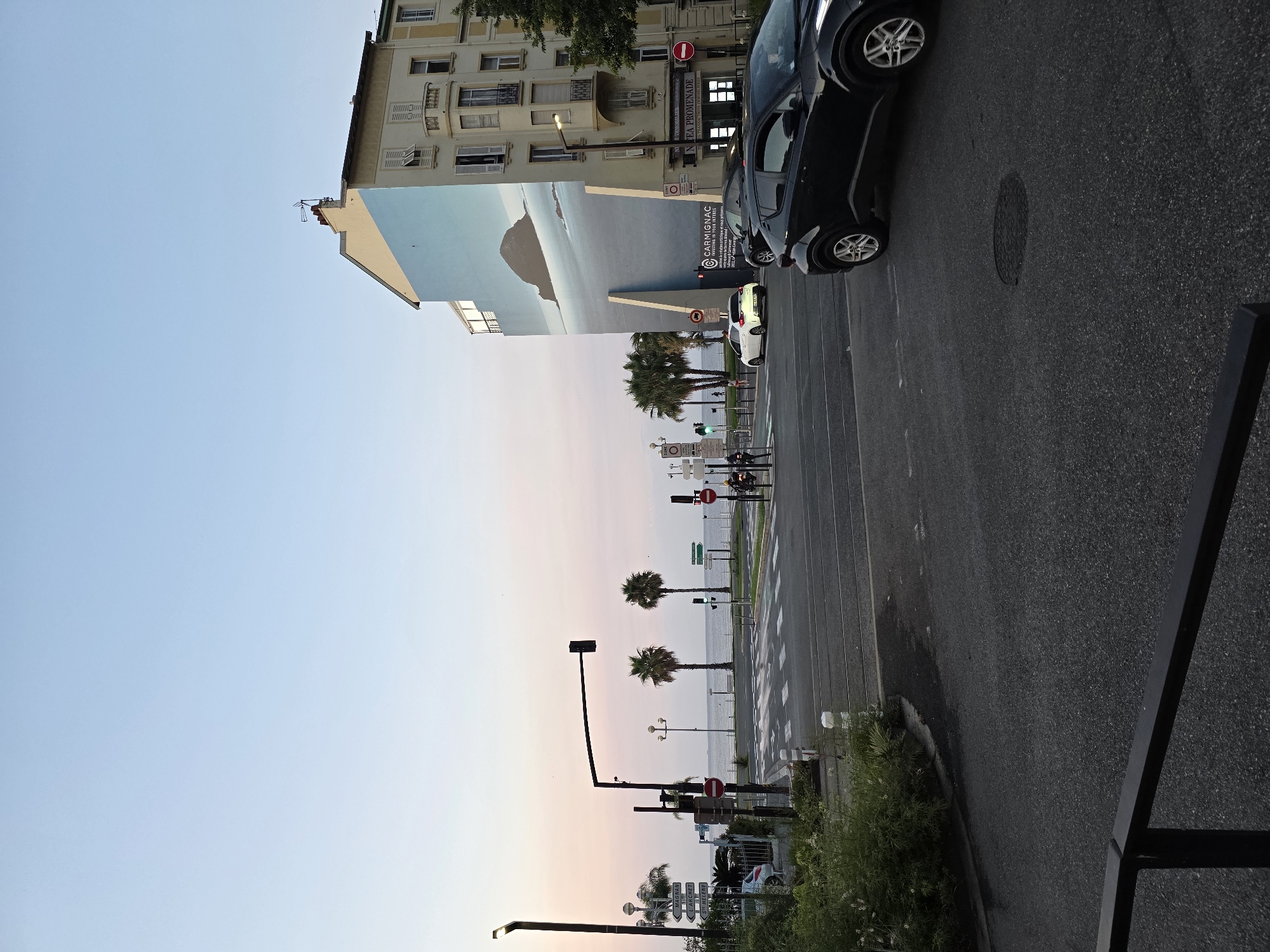 This image depicts a street scene in Nice, France, near the Promenade des Anglais. The time of day appears to be either sunrise or sunset, indicated by the soft, pastel colors in the sky. In the foreground, a paved road with white lane markings and a pedestrian crossing is visible. Two cars are present on the road: a dark-colored SUV in the right lane and a white hatchback turning left in the intersection.
Dominating the right side of the image is a multi-story building with traditional French architecture, featuring light-colored facades and numerous windows with shutters. A large mural of a coastal landscape with a rocky outcrop and water covers a significant portion of this building's side. Streetlights and traffic signals are positioned along the road.
To the left, palm trees line the street, and beyond them, the Mediterranean Sea is visible. Road signs with directional information are mounted on poles. A few people are sparsely distributed on the sidewalk and near the crosswalk. The overall atmosphere is calm and urban, with the natural beauty of the coastline juxtaposed with the built environment.