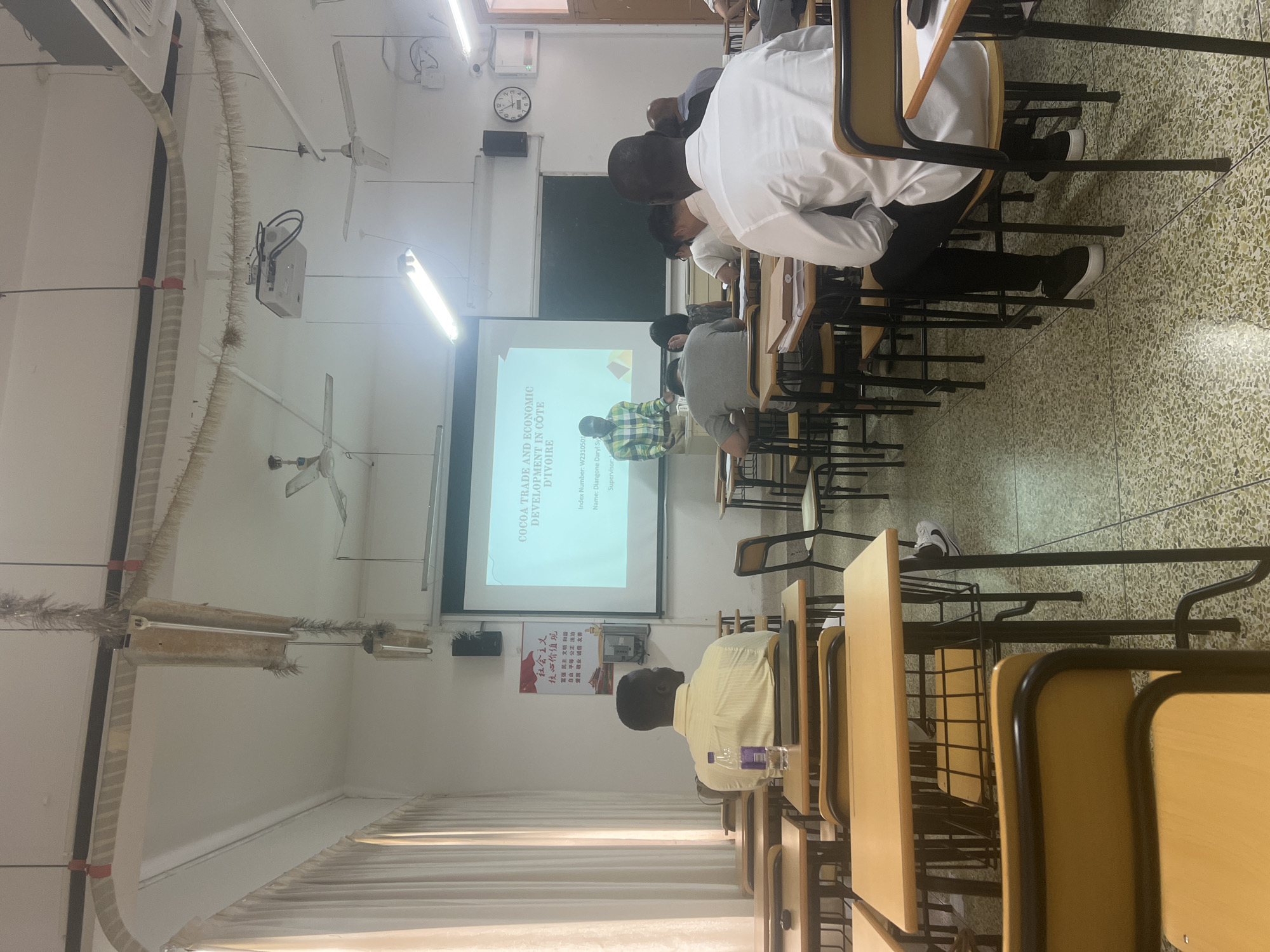This media file captures a classroom scene in what appears to be a lecture or presentation. Several individuals, predominantly men, are seated at desks, facing a projector screen. The screen displays a title slide in English: "COCOA TRADE AND ECONOMIC DEVELOPMENT IN CÔTE D'IVOIRE." Below this, there is text indicating an "Index Number: W2310501," "Name: Diangone Daryl Sy," and "Supervisor:". To the left of this slide, there is a slide with Chinese text, which translates to "Socialist Core Values" followed by a list of phrases.
The room has a typical lecture hall setup with rows of desks and chairs. Overhead lights and ceiling fans are visible, suggesting an indoor environment. The floor has a speckled pattern. The overall atmosphere seems to be one of academic engagement, with individuals focused on the presentation. The people are dressed in casual to semi-formal attire, including shirts and trousers. The lighting suggests it is daytime. The presence of the Chinese text on one of the slides and the provided location context indicates the setting is Liandu District, China.