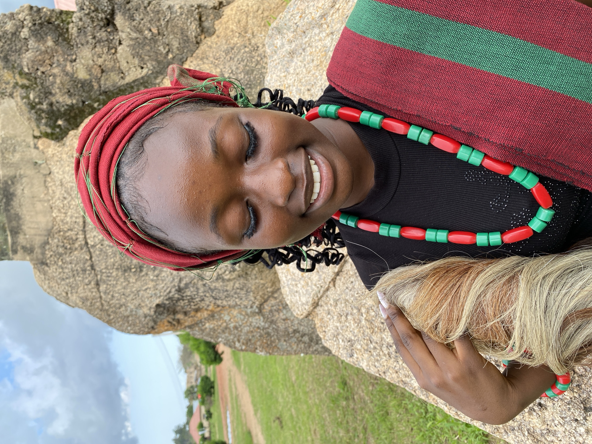The media file shows a young Black woman with her eyes closed and a smile on her face, leaning against a large rock. She is wearing a red headwrap adorned with thin green threads, a black top, and a beaded necklace made of alternating red and green beads. A red and green striped cloth is draped over her shoulder. Her right hand is holding a bundle of light-colored hair. The scene appears to be outdoors, with a grassy field and some trees visible in the background. The sky is cloudy. The image is oriented vertically. This appears to be a portrait of the woman in a natural setting, possibly in Wang, Nigeria. The lighting suggests it is daytime.