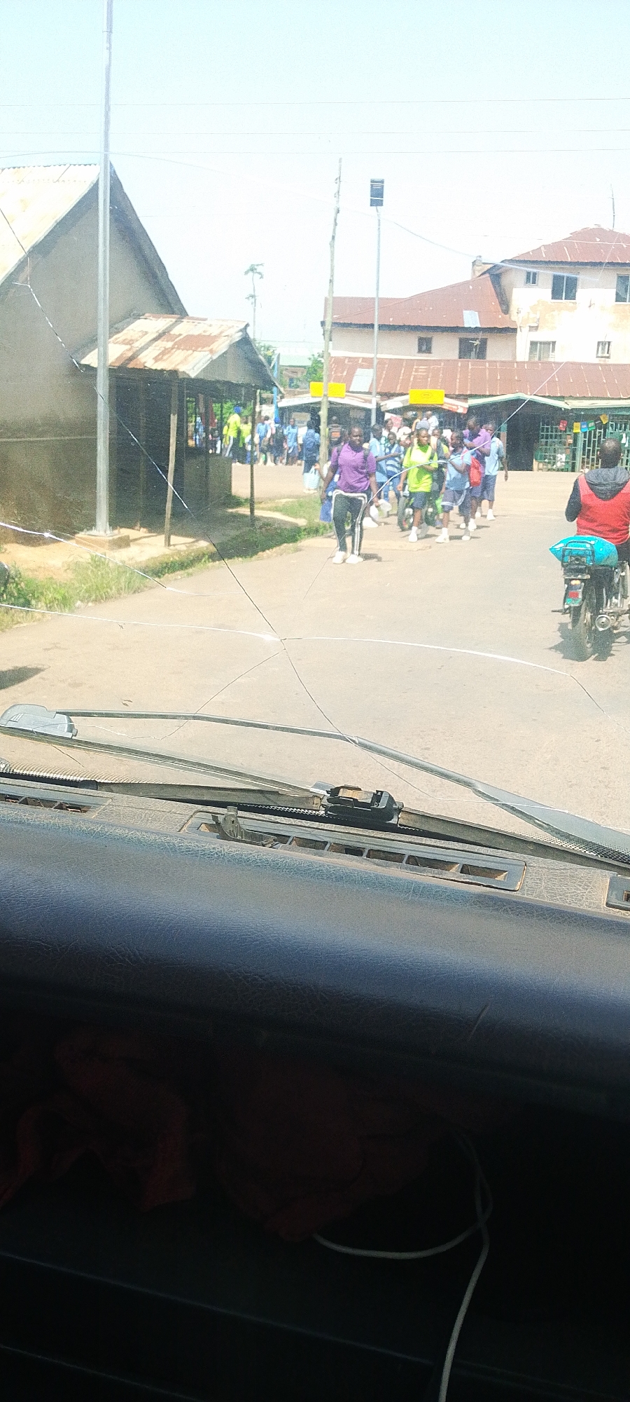 A group of people, some wearing purple and green shirts and blue shorts, walk along a dirt road in front of buildings with corrugated metal roofs. A person wearing a red shirt rides a motorcycle with a blue bag on the back in the foreground. The image is taken from inside a vehicle, with the cracked windshield and dashboard visible. The sky is bright and clear.