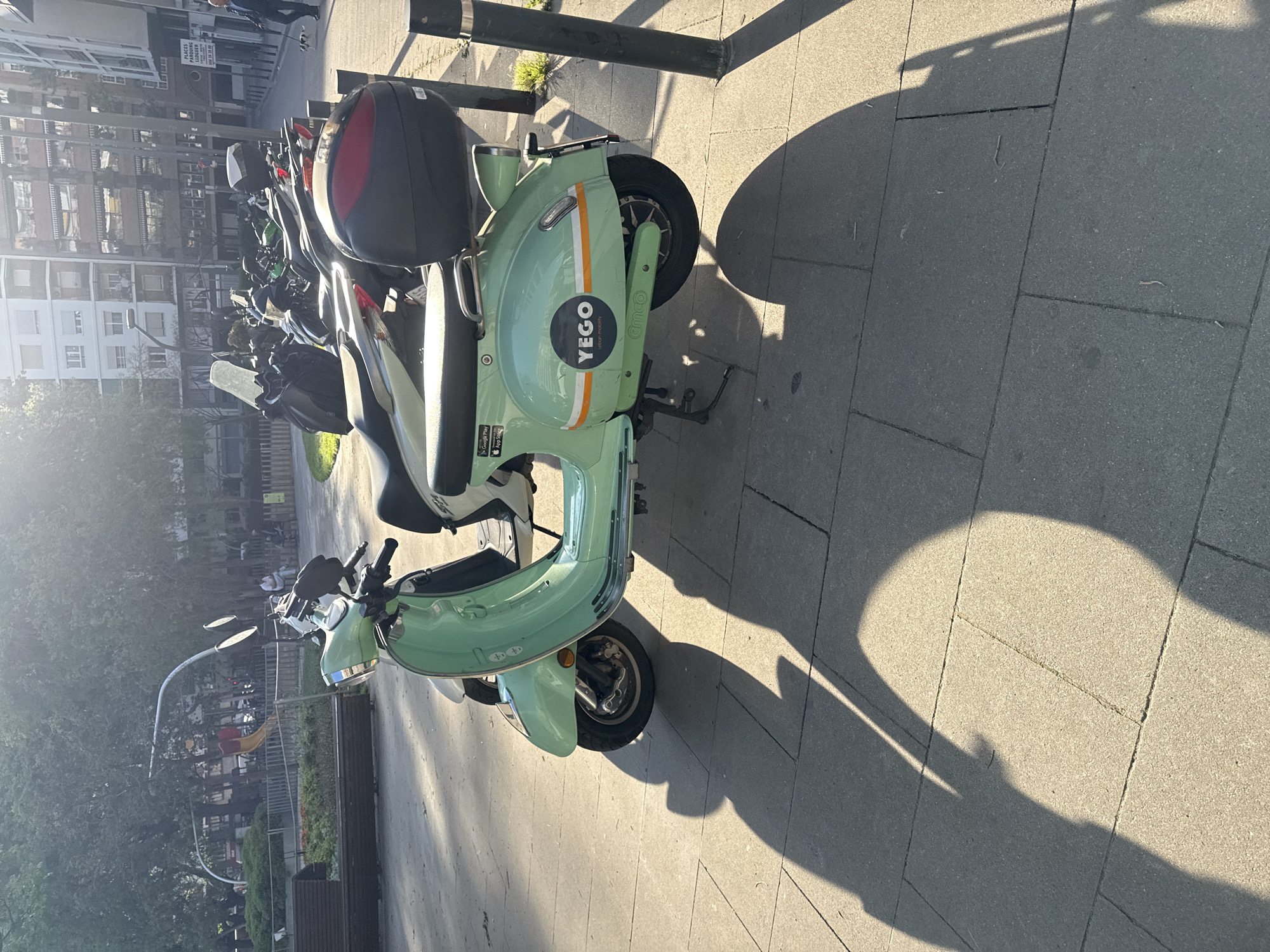 The image shows a light green Yego electric scooter parked on a paved sidewalk in Barcelona, Spain. The scooter is parked at an angle, facing away from the camera. Behind it are other parked scooters. A dark grey storage box is attached to the rear of the scooter. The Yego logo is visible on the side. The sidewalk is grey and the shadow of the scooter is cast on the ground. A metal pole is visible in the background. Across the sidewalk, trees line a park. Buildings and street lamps can be seen in the background.