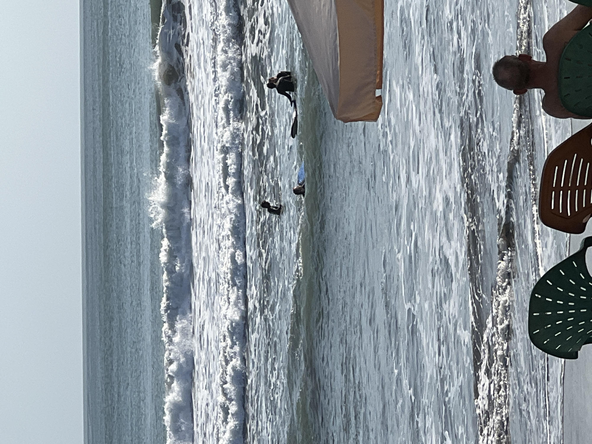 The image depicts a beach scene in Kuta, Indonesia. The ocean is active with moderate waves breaking, and foam churns on the water's surface. Several individuals are in the water, some appearing to be surfing or preparing to surf. One person is lying on a surfboard, while others are standing or wading. In the foreground, two chairs are visible on the sandy shore, with one occupied by a person facing the ocean. A canopy or umbrella provides shade in the mid-ground, partially obscuring the view of the water and surfers. The sky is clear and bright, suggesting a sunny day.