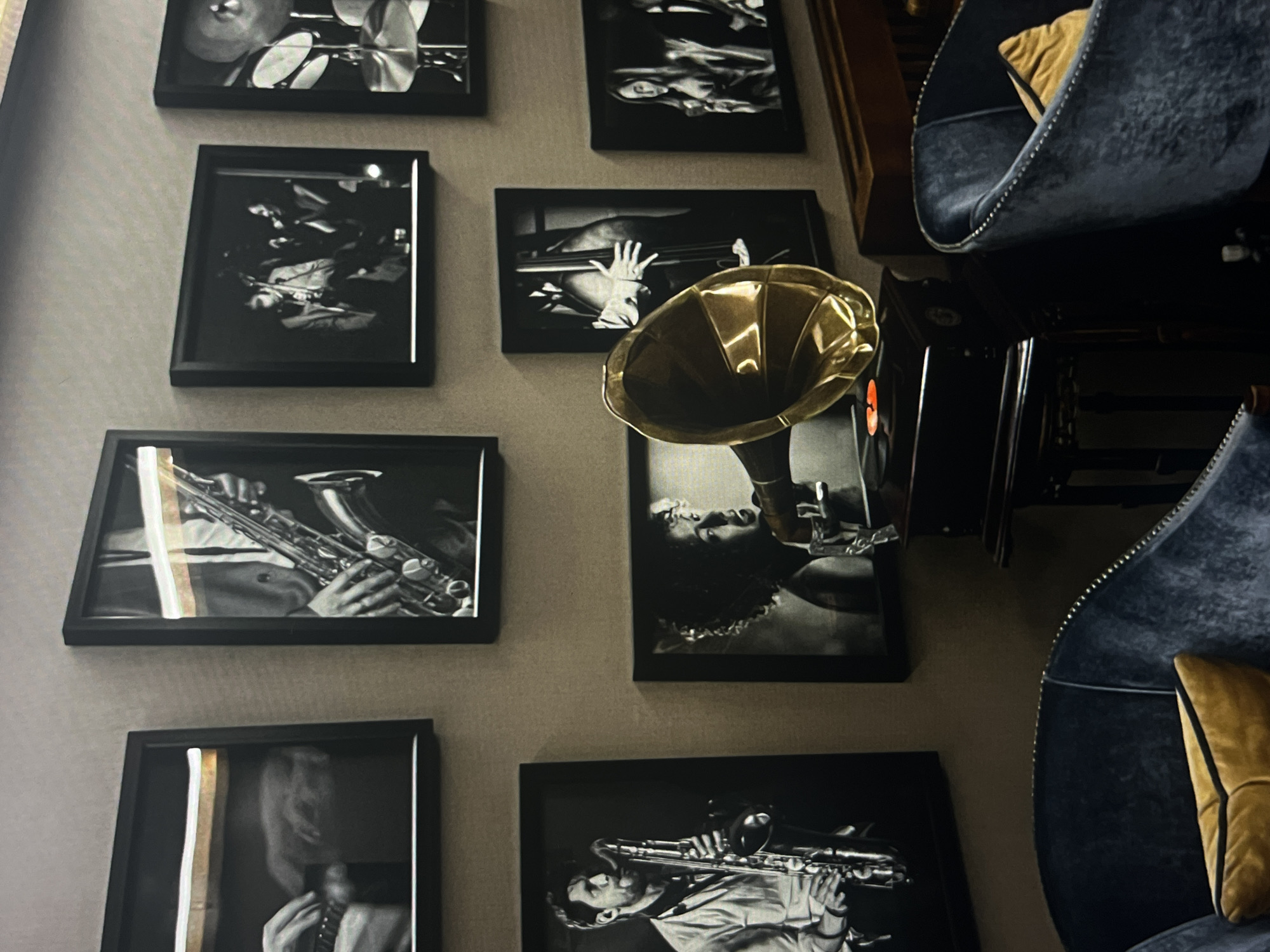 A room with a wall displaying framed black and white photographs of jazz musicians and musical instruments. A golden gramophone sits on a dark surface, possibly a table or shelf, in front of one of the photographs. Two blue velvet chairs with yellow pillows are positioned on the right side of the frame, facing away from the camera. The location in the city is unknown.