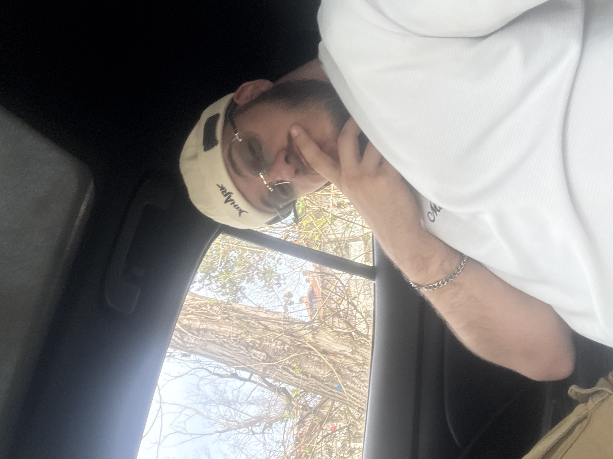 A man is seated inside a vehicle, partially facing the camera. He is wearing a light-colored baseball cap, sunglasses, and a white long-sleeved shirt. A silver chain bracelet is visible on his wrist. His right hand is near his face, with his fingers touching his forehead. The vehicle interior includes a dark-colored dashboard and a grab handle. The view through the window shows a large tree with bare branches. In the background, possibly beyond the tree, are figures of two people positioned high up, suggesting they might be engaged in tree maintenance. The location of the scene is Teteven, Bulgaria.