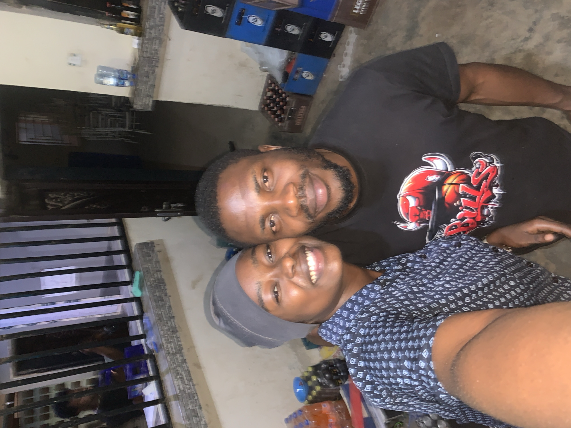 Two individuals are posing for a photo. One individual is wearing a black t-shirt with a red graphic on the front. The other individual is wearing a patterned blue shirt and a gray head covering. The background contains stacks of beer crates, bottles of water, and a window with bars. The location appears to be in or near a residential building, potentially in the city of New Stadium, Nigeria.