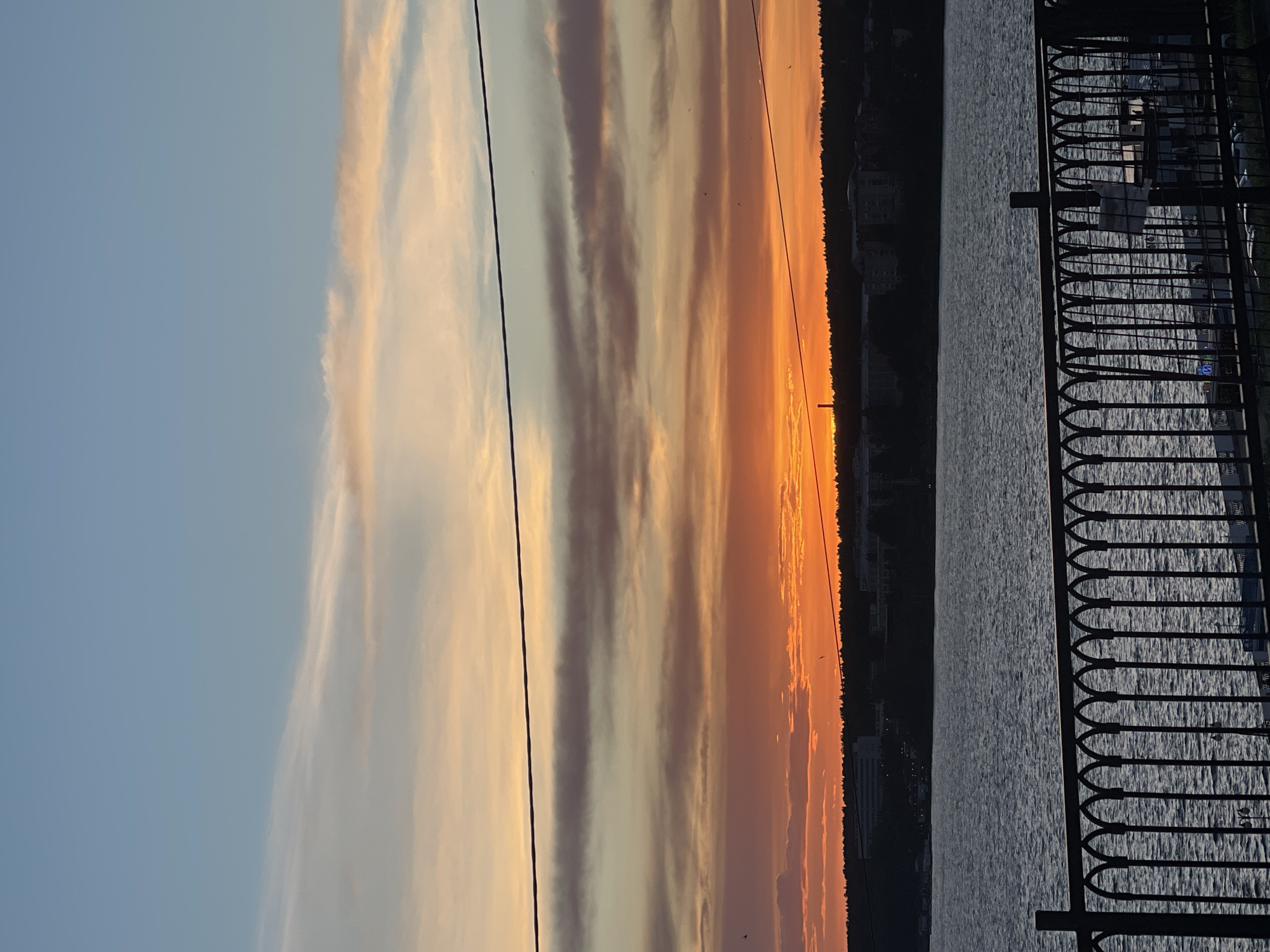 The image shows a sunset over a body of water, possibly a river or lake, as seen from a vantage point behind a decorative metal fence. The sky exhibits a gradient of colors, ranging from light blue to shades of orange and red near the horizon. Long, streaky clouds are present in the sky. A line of trees or a cityscape is silhouetted against the sunset. The water is dark, reflecting the light from the sky. Two dark wires run vertically through the image. The location is most likely a riverside or lakeside area in a city.