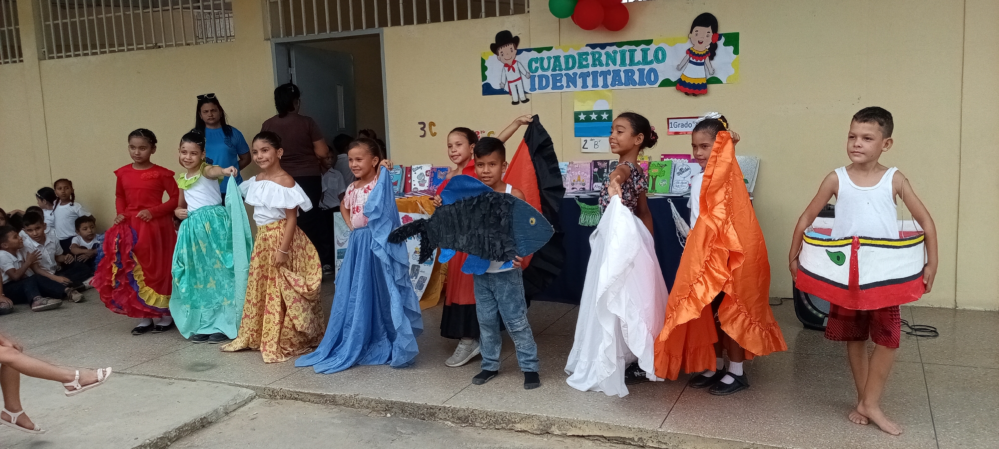 The image shows a group of children standing on a concrete surface, likely a stage or elevated platform, in front of a building. The building has a beige exterior with a doorway visible in the background. Behind the children is a banner that reads "CUADERNILLO IDENTITARIO" and has illustrations of people. There are also posters displayed on the wall above a table covered with a dark blue cloth.
The children are dressed in various colorful costumes and carrying props. A boy in the center is holding a large, blue, fish-shaped cutout. Some children are holding pieces of fabric in different colors, such as white, orange, and black. One child is wearing a costume that resembles a boat.
In the background, there are additional people sitting and standing, including adults who appear to be supervising the children. The overall scene appears to be a school event or cultural presentation. The city and specific location are unknown from the image alone.