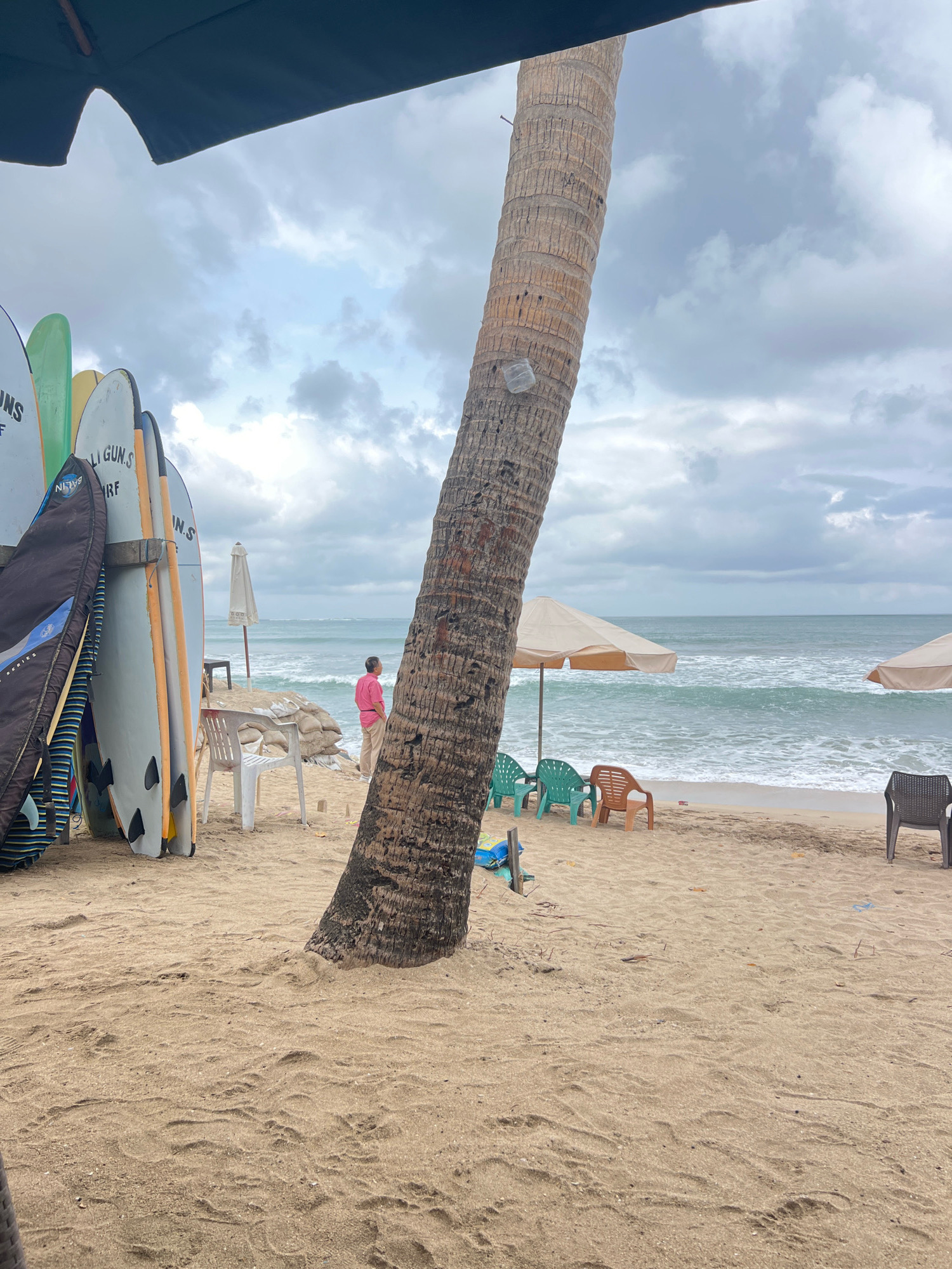 The image captures a scene on the beach in Kuta, Indonesia, under a cloudy sky. In the foreground, a large palm tree trunk is prominent on the sandy beach, with its base partially obscured by the sand. To the left of the palm tree, several surfboards are propped up. They are of various colors and designs, including some with visible logos or text. A black surfboard bag is also present.
Further into the scene, on the sand, are several beach chairs and umbrellas. To the left, a white plastic chair is positioned near a few sandbags and a small white umbrella. To the right, there are several brightly colored plastic chairs – two green and one orange. Further to the right, a dark-colored chair is visible.
In the background, the ocean stretches out with moderate waves rolling towards the shore. The horizon is visible, with the sea meeting the sky. A person wearing a pink shirt and beige pants is standing on the beach, facing the ocean, looking out at the water. The overall environment suggests a typical beach setting in Kuta, a popular surfing destination.