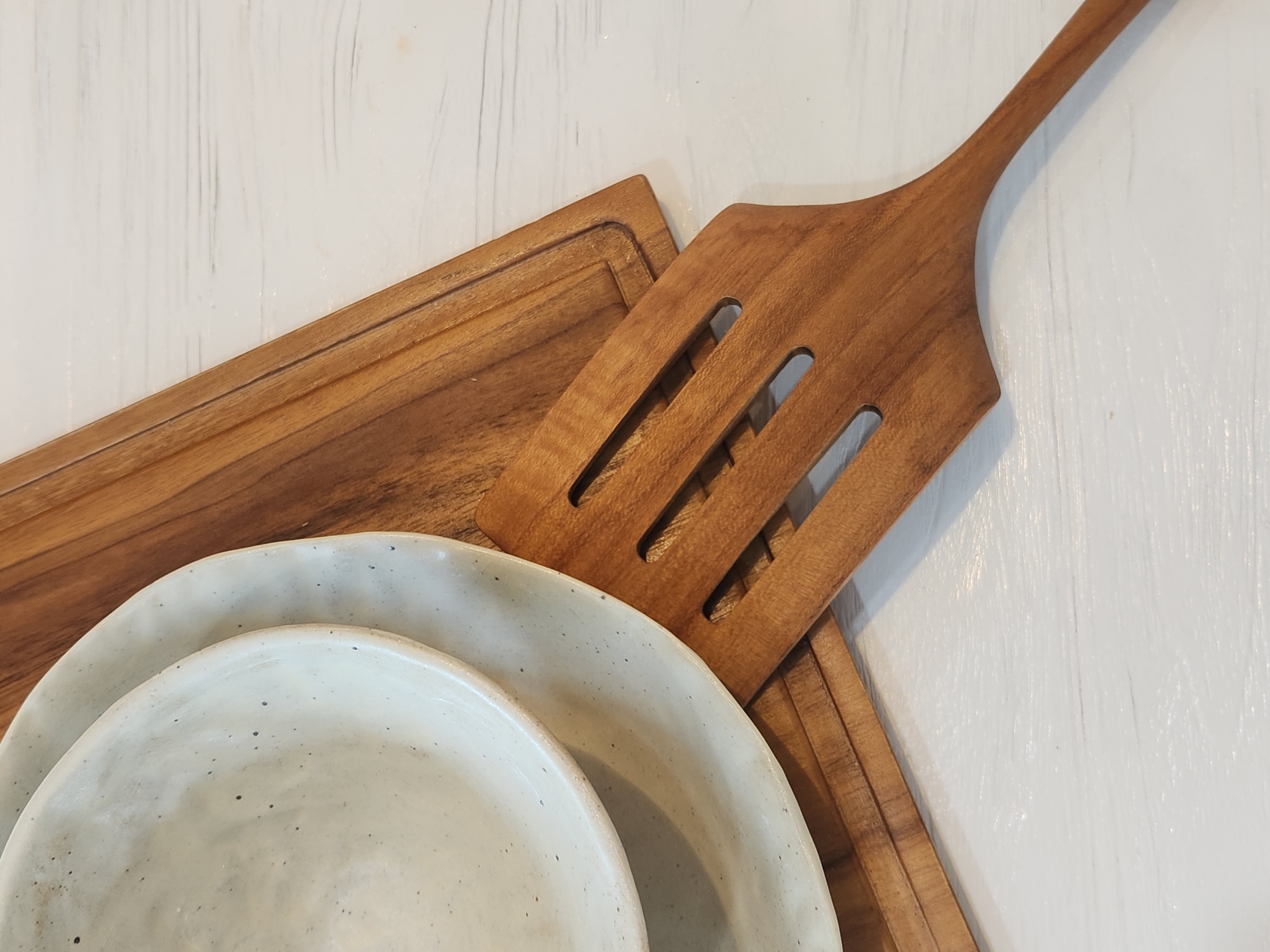 The image shows a close-up, overhead view of kitchenware arranged on a white, textured surface. The primary objects are two off-white ceramic bowls, one nested inside the other, and a wooden spatula. The spatula is made of a warm-toned wood and has several rectangular slots in its wider end, suggesting it's a serving or cooking utensil. It lies diagonally across the frame, with its handle extending towards the top right corner.
Beneath the spatula, partially visible, is a rectangular wooden cutting board. The cutting board has a subtle groove around its perimeter. The ceramic bowls are positioned in the lower left quadrant of the image. They have a speckled, handmade appearance, with visible texture and small dark flecks against the off-white glaze.
The white surface beneath the objects appears to be painted wood, with visible brushstrokes that create a slightly irregular texture. The lighting is soft and diffused, casting no harsh shadows, and suggests an indoor setting, possibly a kitchen or dining area. There is no indication of a specific time of day, weather, or any discernible emotions. No text is visible in the image. The overall aesthetic is rustic and simple, highlighting the natural materials of the wood and ceramic.