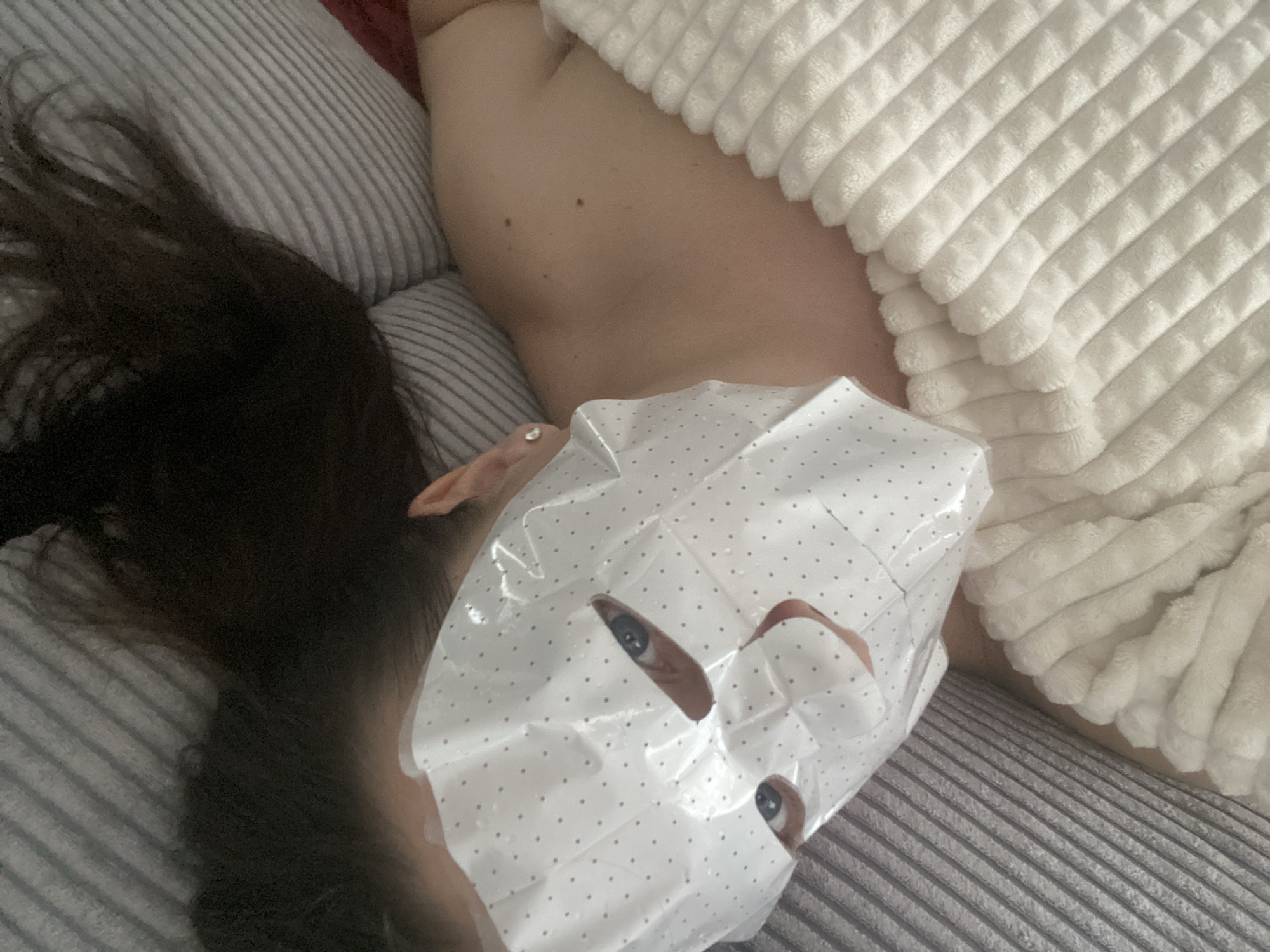 The photograph depicts a person lying down with a face mask on. The person's head is positioned on a gray striped pillow, and their dark hair is visible to the left. Part of the person's body is covered by a white, textured blanket. The face mask has a white background with small gray dots and cutouts for the eyes and mouth. The person's eyes are visible through the mask. No information is available regarding the location of the city of Sochi, Russia, or any activity.