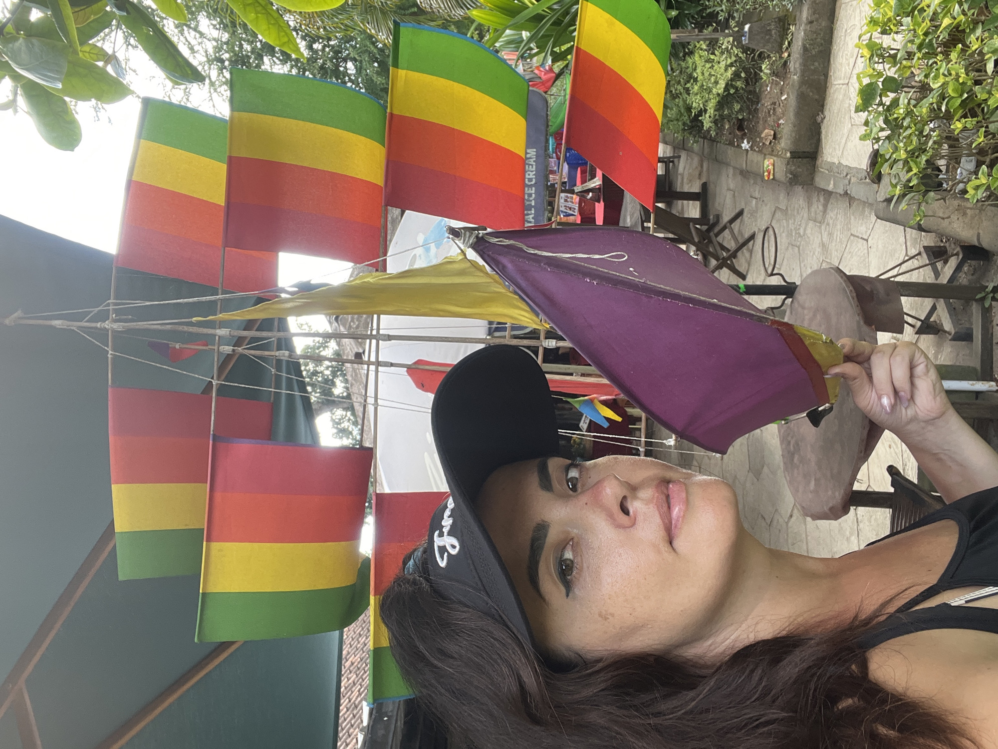 The image shows a woman in the foreground holding a small purple kite, with a portion of a black cap on her head that has the word "June" embroidered in white. Behind her, a structure resembling a ship's mast is visible, adorned with several rainbow-colored flags, each divided into vertical stripes of green, yellow, red, and a dark pink or purple. The flags are suspended by ropes. The background suggests an outdoor setting with plants and what appears to be an establishment with tables and chairs. A sign partially visible on one of the sails reads "TAL ICE CREAM." The overall scene suggests a recreational or tourist area. Based on the presence of the rainbow flags often associated with the LGBTQ+ community, and the tropical foliage, this location could be a cafe or shop catering to tourists in Kuta Selatan, Indonesia, an area known for its beaches and vibrant atmosphere.