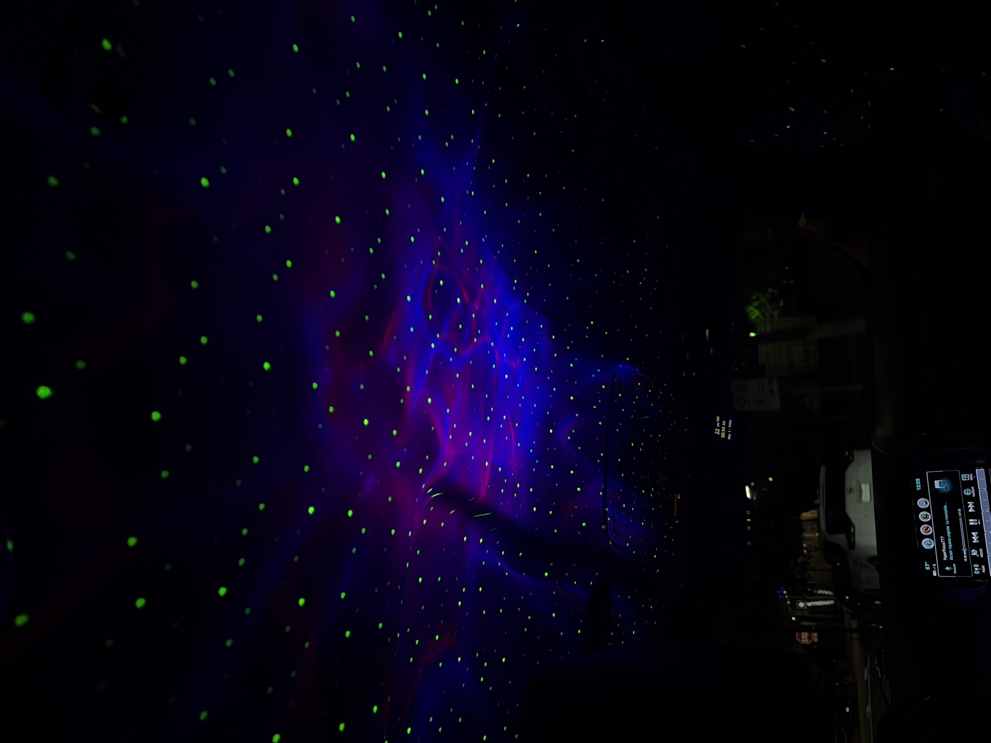 The media file captures the interior of a vehicle at night, illuminated by a dynamic light projector that casts green dots and swirling purple and blue nebula-like patterns across the ceiling and upper panels. The scene is primarily dark, with the vibrant light display creating a cosmic atmosphere within the car.
On the right side of the image, a car's infotainment system is visible, displaying various pieces of information. The time "12:35" is shown, along with a temperature reading of "67°". Music playback details are present, indicating "HyperFocus-777" and "Ahorra espacio urgente. La man..." (likely "manana" or similar) by an unknown artist, with a progress bar showing "1/7:20". Icons for Bluetooth, Radio, and Video are also discernible, along with music control buttons (play/pause, skip, volume).
Looking out through the vehicle's window on the right, a dark street scene is visible, suggesting the car is either parked or driving at night. Distant lights and silhouettes of buildings or trees can be faintly seen. Visible text on a street sign through the window includes "STOP" and partially, "WYNDHAM". Additionally, reflected on an upper part of the dashboard or mirror, some text reads "22 - Nov", "00:54 AM", and "Mon | Happy".
The overall impression is of a car interior transformed by special lighting, possibly for a relaxed drive or a unique ambiance, while parked in Austin, United States of America.
