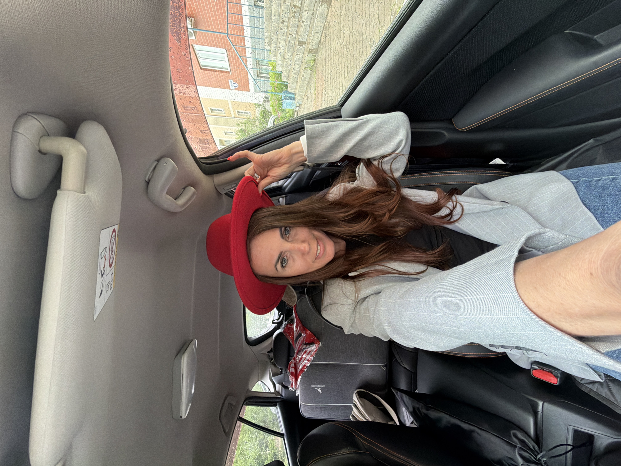 A woman is captured in a sideways-oriented selfie, sitting in the passenger seat of a car. She is smiling directly at the camera, showcasing her long, wavy brown hair, blue eyes, and a bright red wide-brimmed hat. She is dressed in a light grey pinstriped blazer, a dark top, and blue jeans. Her left arm, holding the camera, is partially visible, while her right hand is positioned near her hat, possibly adjusting it or making a slight gesture.
The car's interior features grey and black upholstery with some orange stitching visible on the seats or door panels. A grey sun visor with a safety warning label (featuring an airbag symbol and partially legible text like "AIRBAG") is visible above her head. Through the car windows, numerous raindrops are apparent, indicating rainy conditions. Outside, multi-story brick buildings with windows, typical of urban or residential areas, can be seen, providing a glimpse of Birobidzhan, Russia.
On the back seat, a grey backpack with a white stripe and a logo that appears to be a kangaroo or similar animal is visible. Also on the back seat are a red and white striped bag and a black drawstring bag. The image conveys a casual, relaxed moment, with the woman appearing cheerful despite the rainy weather outside.