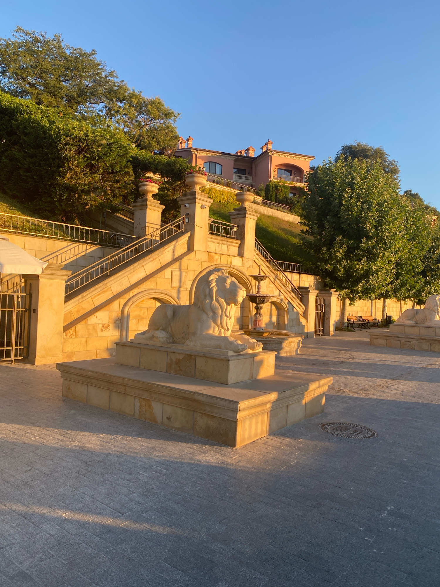 This image depicts an outdoor scene in Odesa, Ukraine, likely during the golden hour as indicated by the warm, low-angled sunlight casting long shadows. In the foreground, a paved plaza with a grey, brick-like pattern leads towards a stone staircase. Flanking the lower section of the stairs are two white, stone lion sculptures, one prominently featured on the left. The stairs themselves are made of light-colored stone blocks and are lined with metal railings.
Behind the stairs, a fountain with multiple tiers is visible. The staircase ascends towards a large, pale pink building situated on a grassy hill. This building features balconies and arched windows, suggesting it might be a hotel, a grand residence, or a public building. Lush green trees and shrubs surround the building and the hillside.
To the right of the main staircase, a similar, though less detailed, stone lion sculpture is also present. Further to the right, a few park benches are visible under a large tree, suggesting a public space or park area. The overall atmosphere is serene and architectural, highlighting the ornate stonework and the natural beauty of the surroundings. There are no people visible in the image.