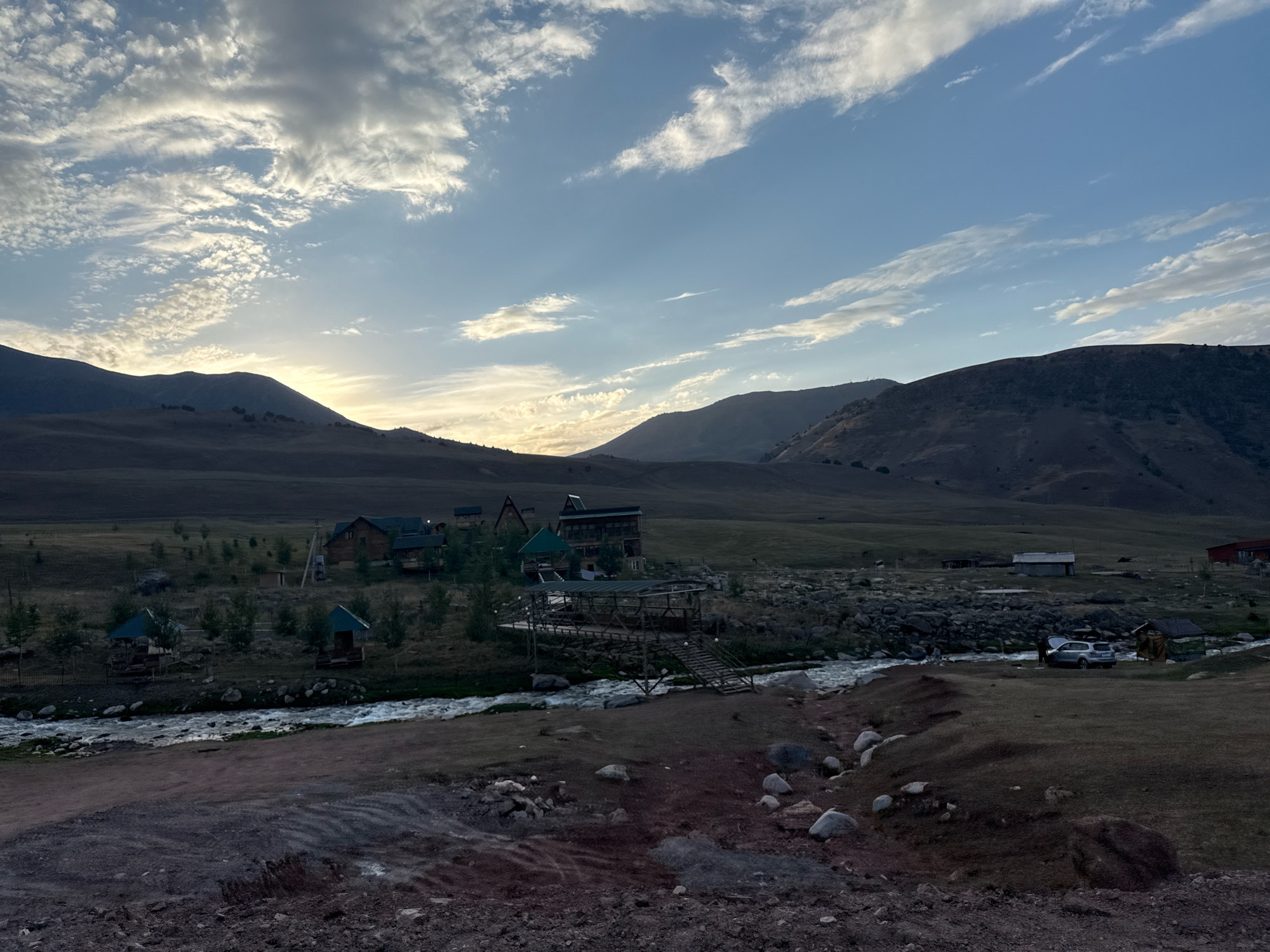 The image depicts a scenic rural landscape in Chunkurchak, Kyrgyzstan, with rolling hills and mountains in the background. The foreground features a winding river with a rocky bed, and a dirt path leading down towards it. On the left bank of the river, there are several small, colorful structures resembling gazebos or shelters, nestled amongst sparse trees. Further inland, a cluster of wooden houses and a larger, multi-story building with a dark roof are visible. A wooden bridge or walkway spans the river, connecting to the buildings. To the right of the river, a grey SUV is parked near a small, dark-colored structure. The sky is partly cloudy with streaks of sunlight breaking through, suggesting either sunrise or sunset. The overall scene conveys a sense of peaceful, natural beauty, with elements of human habitation integrated into the landscape.