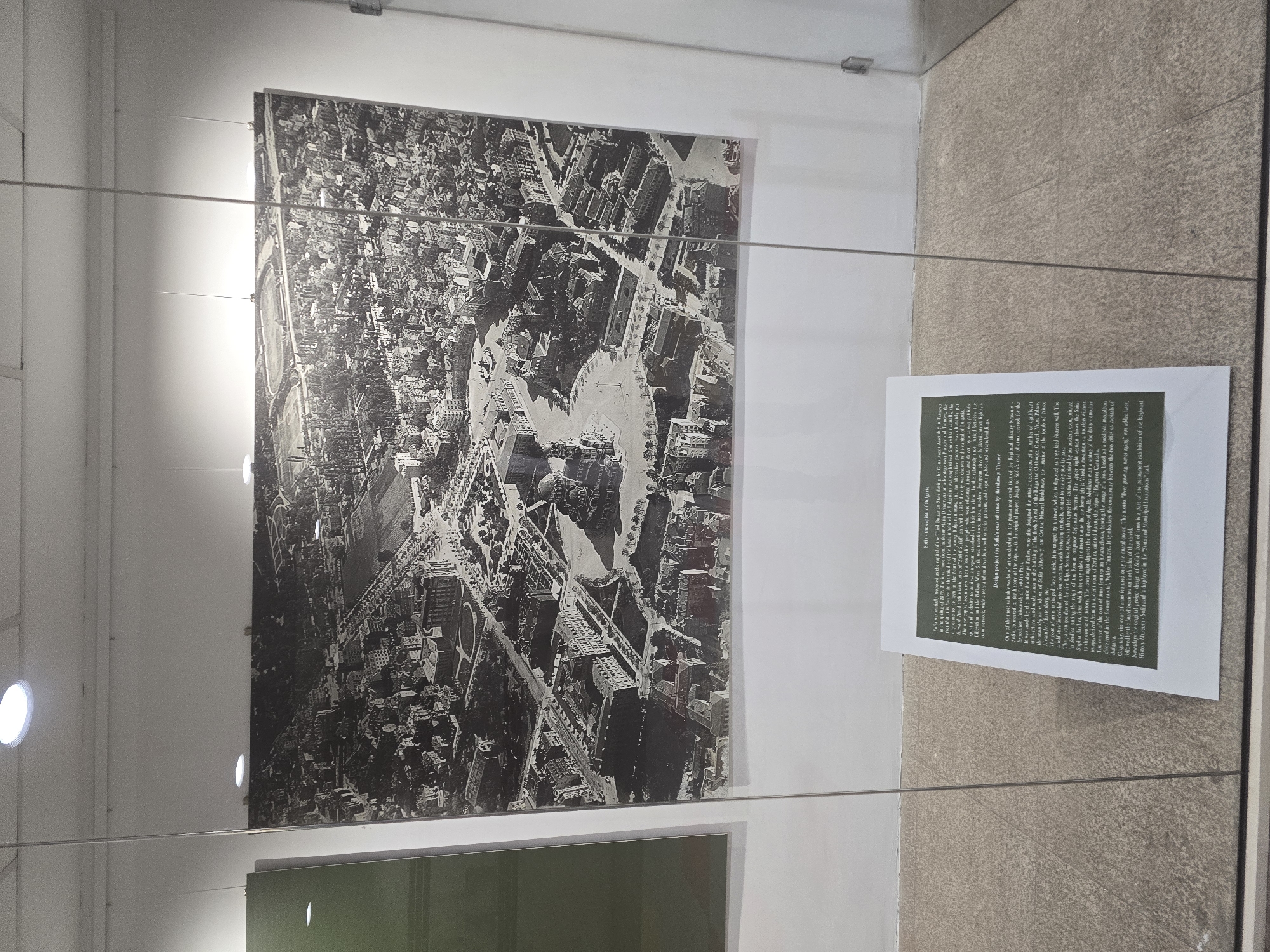 The image shows a display in a museum featuring two framed items: a large black-and-white aerial photograph and a smaller descriptive plaque.
The photograph depicts an aerial view of a city, presumably Sofia, Bulgaria, with numerous buildings and streets visible. The layout shows complex urban planning and architecture of the time.
To the right of the photograph is the framed plaque, which features text in English, describing the history of Sofia and the design of its coat of arms. The text provides historical context for the city and the symbolism within its coat of arms.
The items are displayed in what appears to be a glass-enclosed exhibit, with a light source visible on the left wall illuminating the display. The floor and walls of the exhibit are a neutral beige color. The environment is sterile and well-lit, consistent with a museum setting.