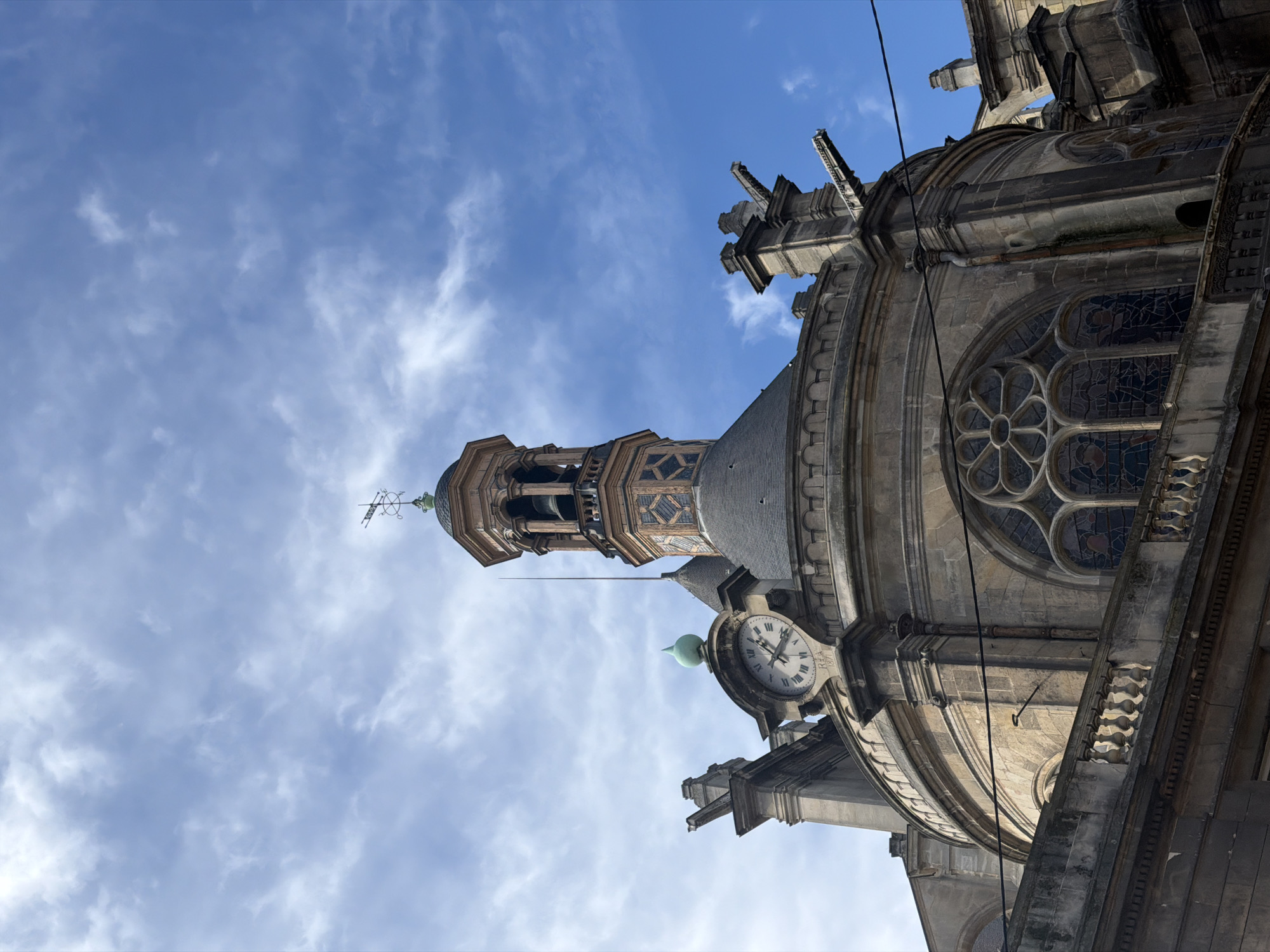 This image displays a detailed view of the upper architecture of the Église Saint-Eustache, situated in the 1st arrondissement of Paris, France. The scene is dominated by a stone building featuring a large, dark-tiled dome from which an octagonal bell tower ascends. The tower is embellished with decorative brickwork, houses a visible bell within its arched openings, and is topped by a smaller dome, a slender lightning rod, and a weather vane marked with cardinal directions. To the left of the central tower, a circular clock face with Roman numerals, indicating approximately 10:45, is integrated into the stone structure. Below the clock, the inscription "R.E.A. 1871" is legible. The building's facade includes multiple stone gargoyles or drainage spouts projecting from upper cornices. Lower sections reveal large arched windows with elaborate stained glass, featuring a rose window pattern and figurative elements, partially obscured by a classical stone balustrade. The stone construction exhibits weathering and dark streaks, consistent with age. Overhead, a bright blue sky with scattered white clouds is visible. Thin black utility lines traverse the mid-section of the building's facade. No people or actions are depicted.